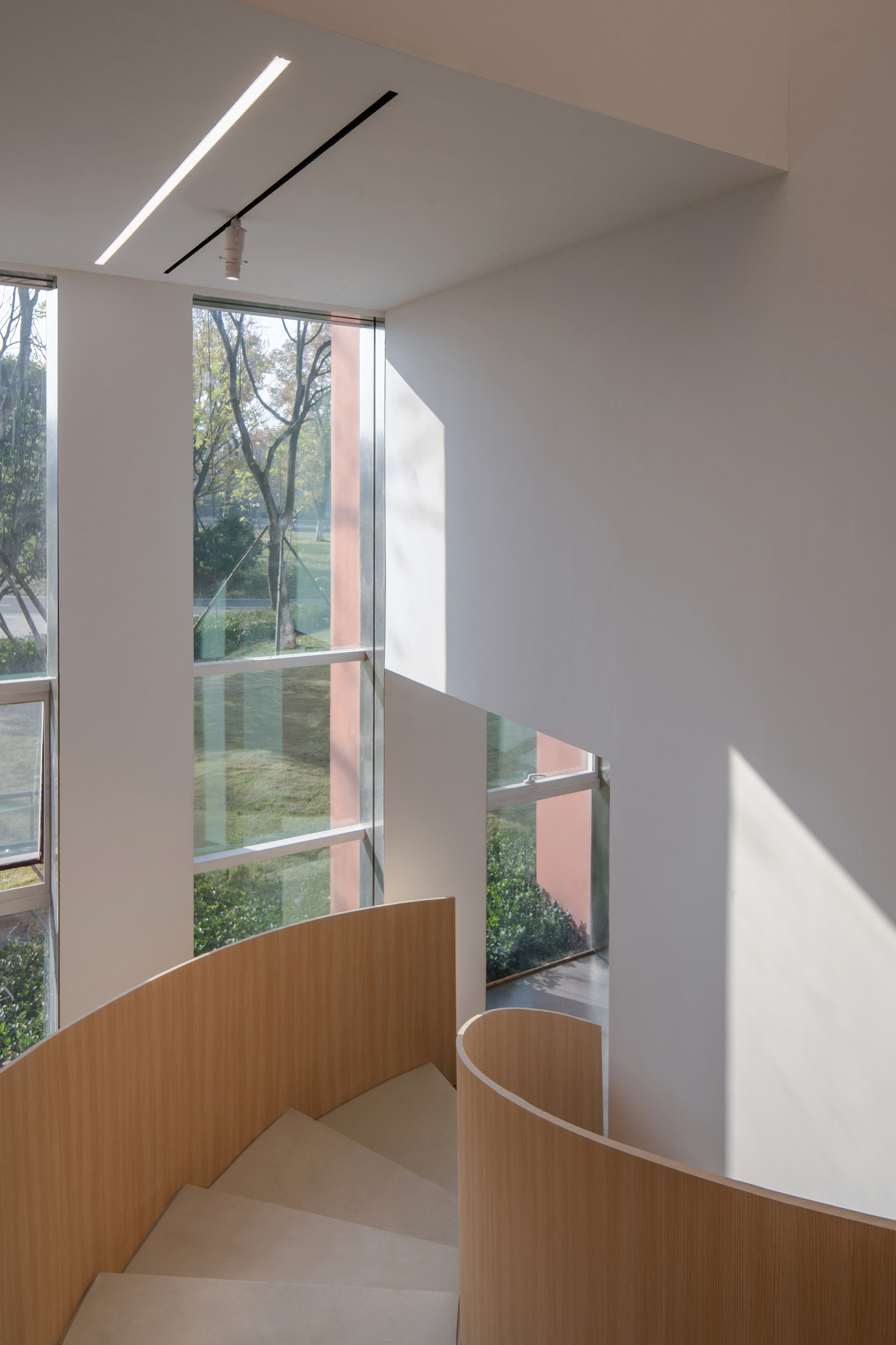 View down the curved wooden stair wall toward tall glazing framing bare trees and a lawn in soft light