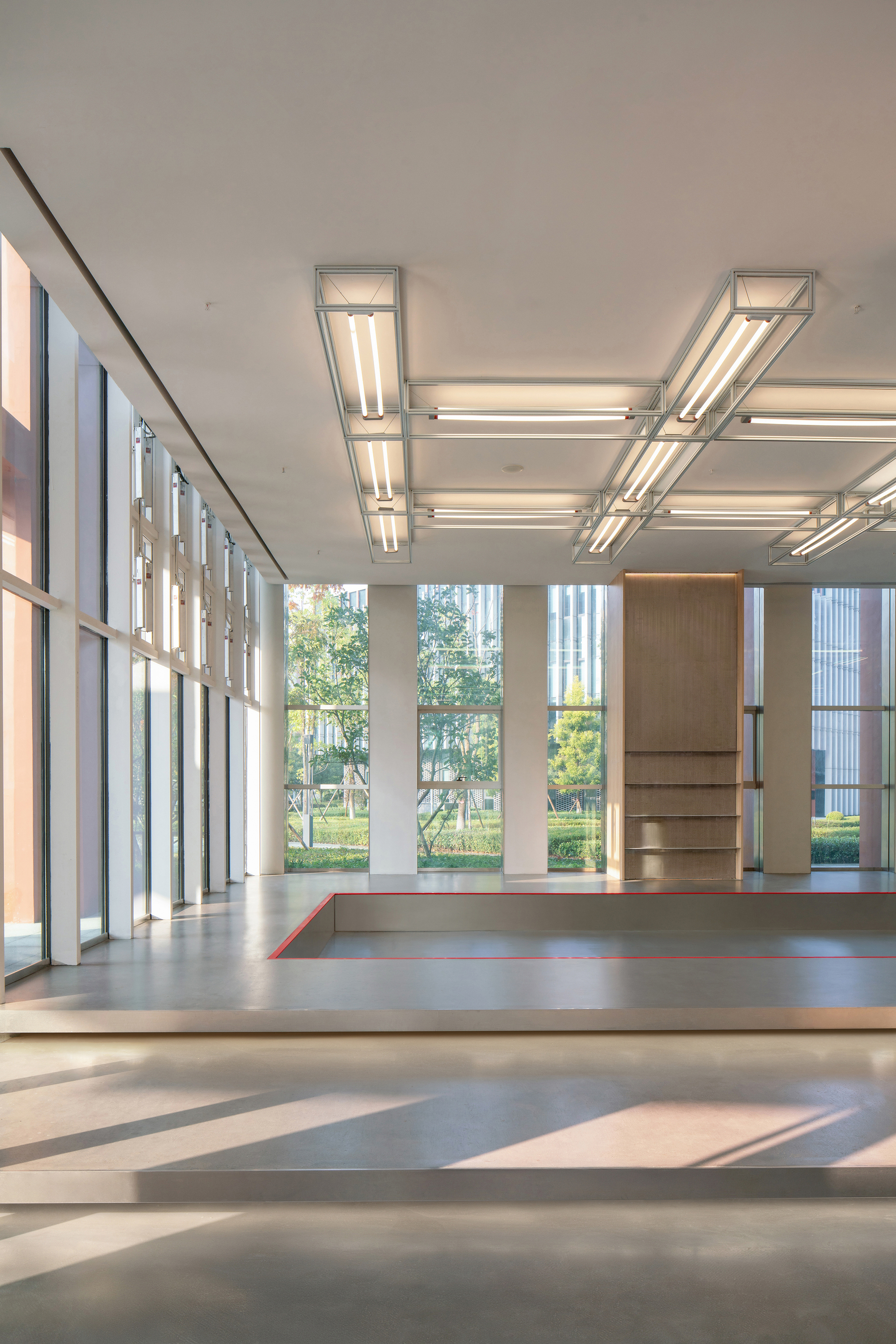 Raised platform with red-edged steps and a timber shelf unit, under steel-framed fluorescent fixtures and afternoon sun through full-height glazing