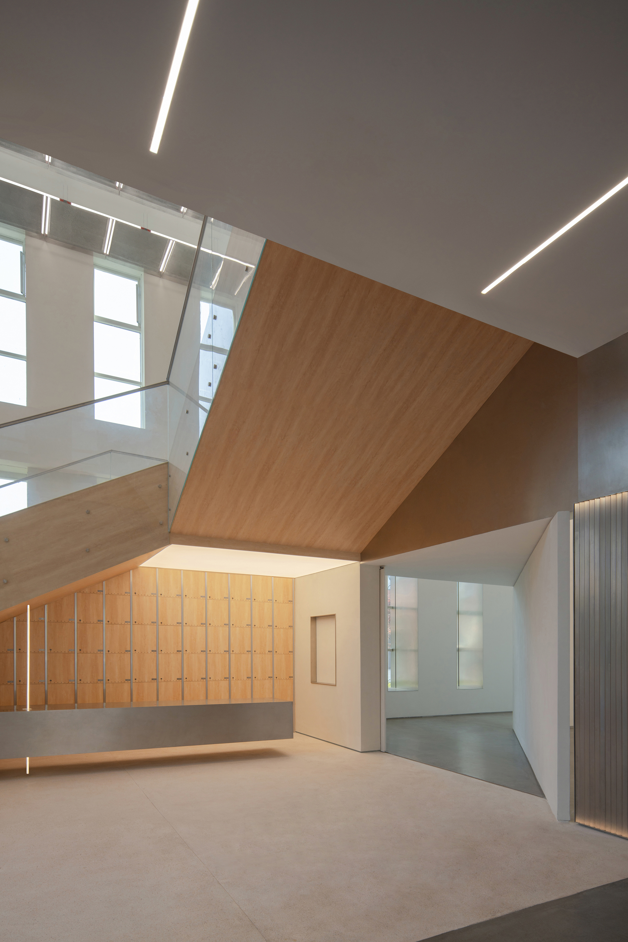 Angled timber-lined ceiling plane above a wall of wooden lockers and a glass balustrade, with linear lights overhead