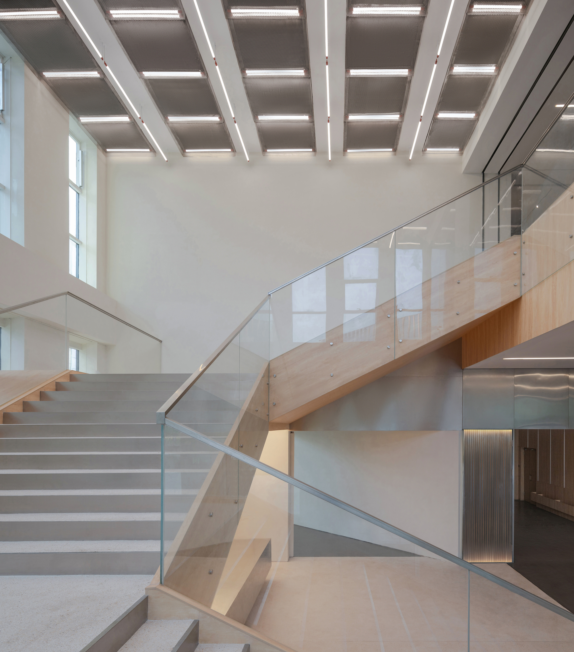 Glass-balustraded stair with timber stringers rising beneath a coffered ceiling of recessed light panels in a tall white atrium