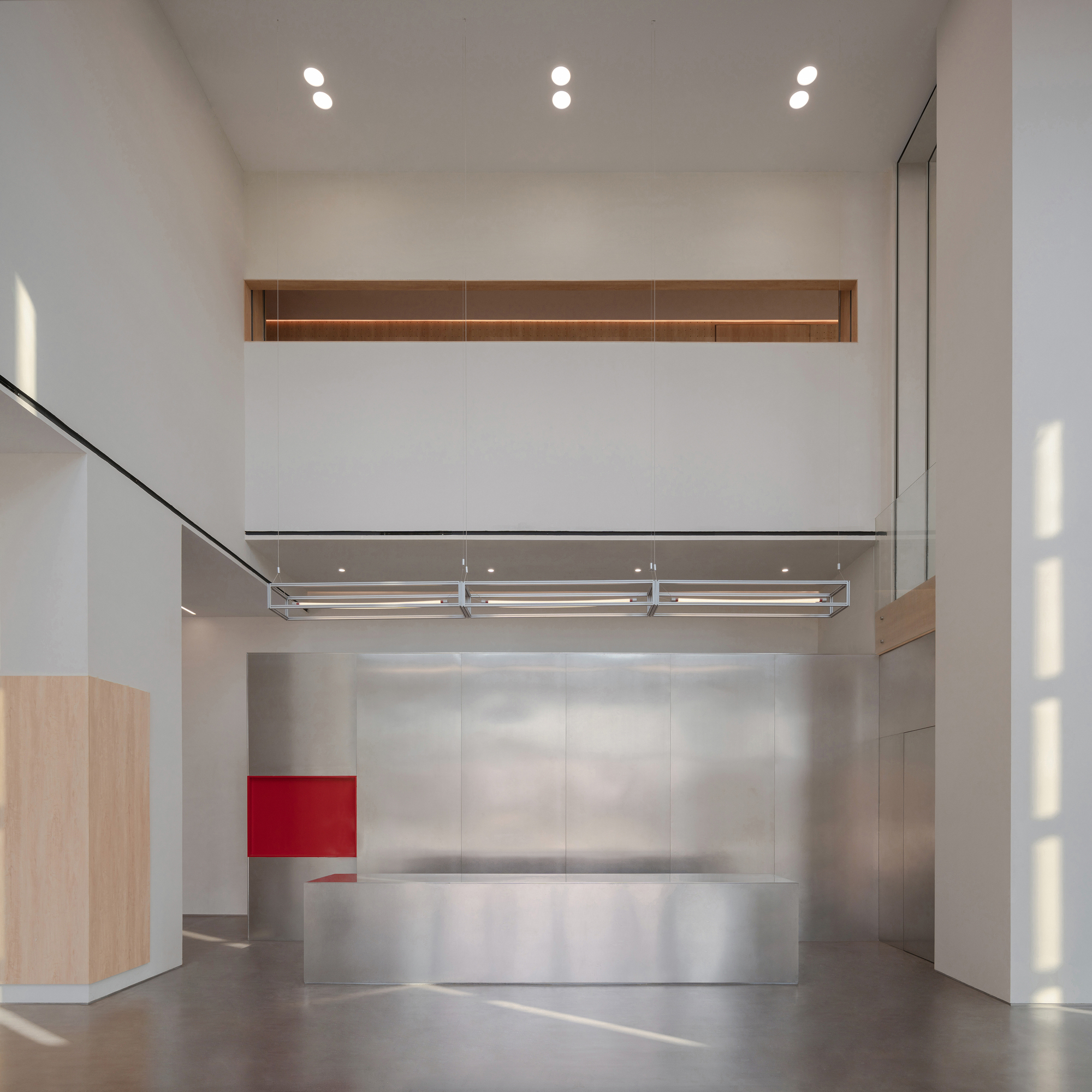 Double-height lobby with a brushed steel reception counter, red panel and suspended light frame under soft daylight