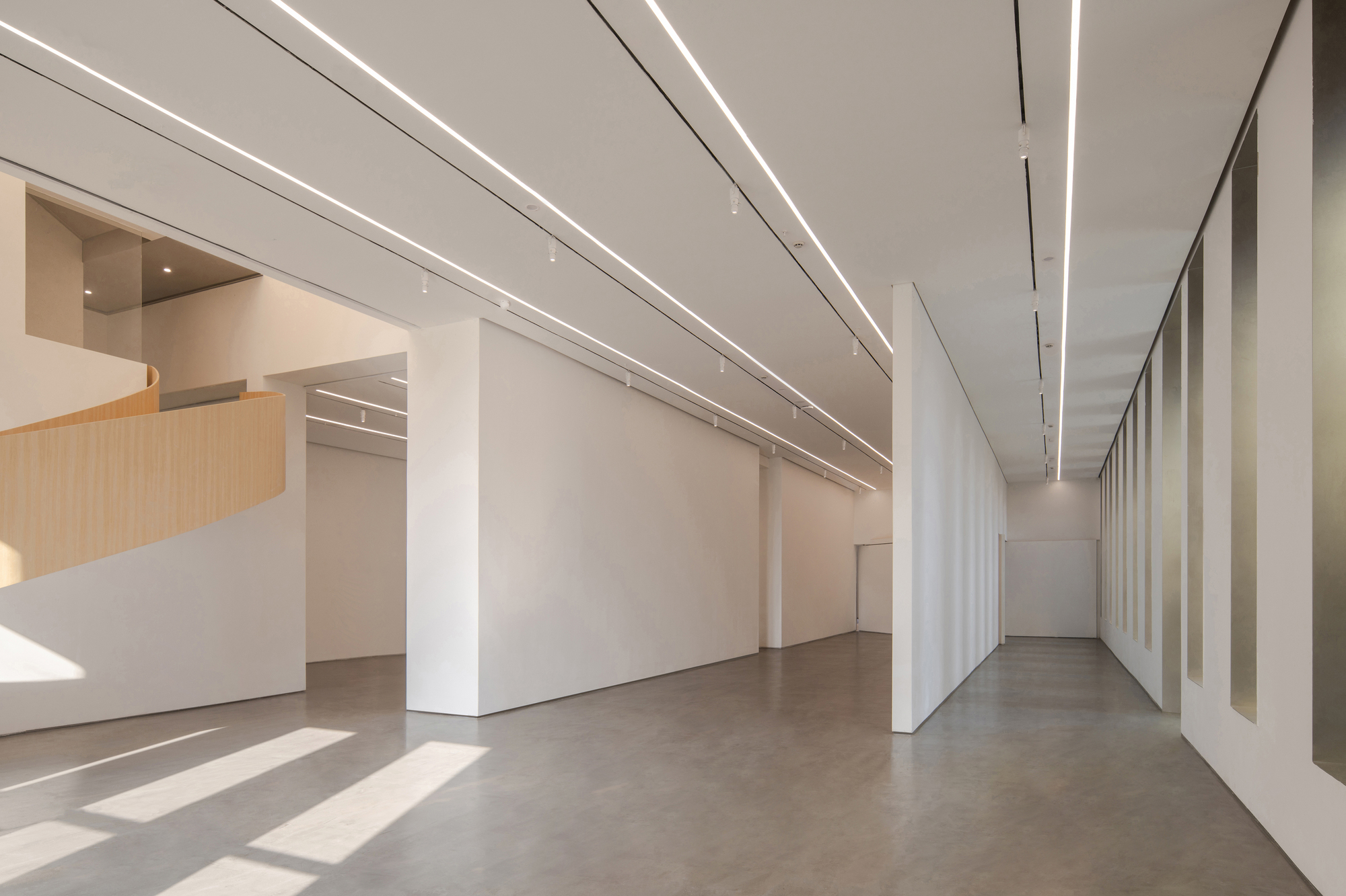 Gallery hall with white partition walls and linear ceiling lights, sun patches on the floor and the timber stair at left