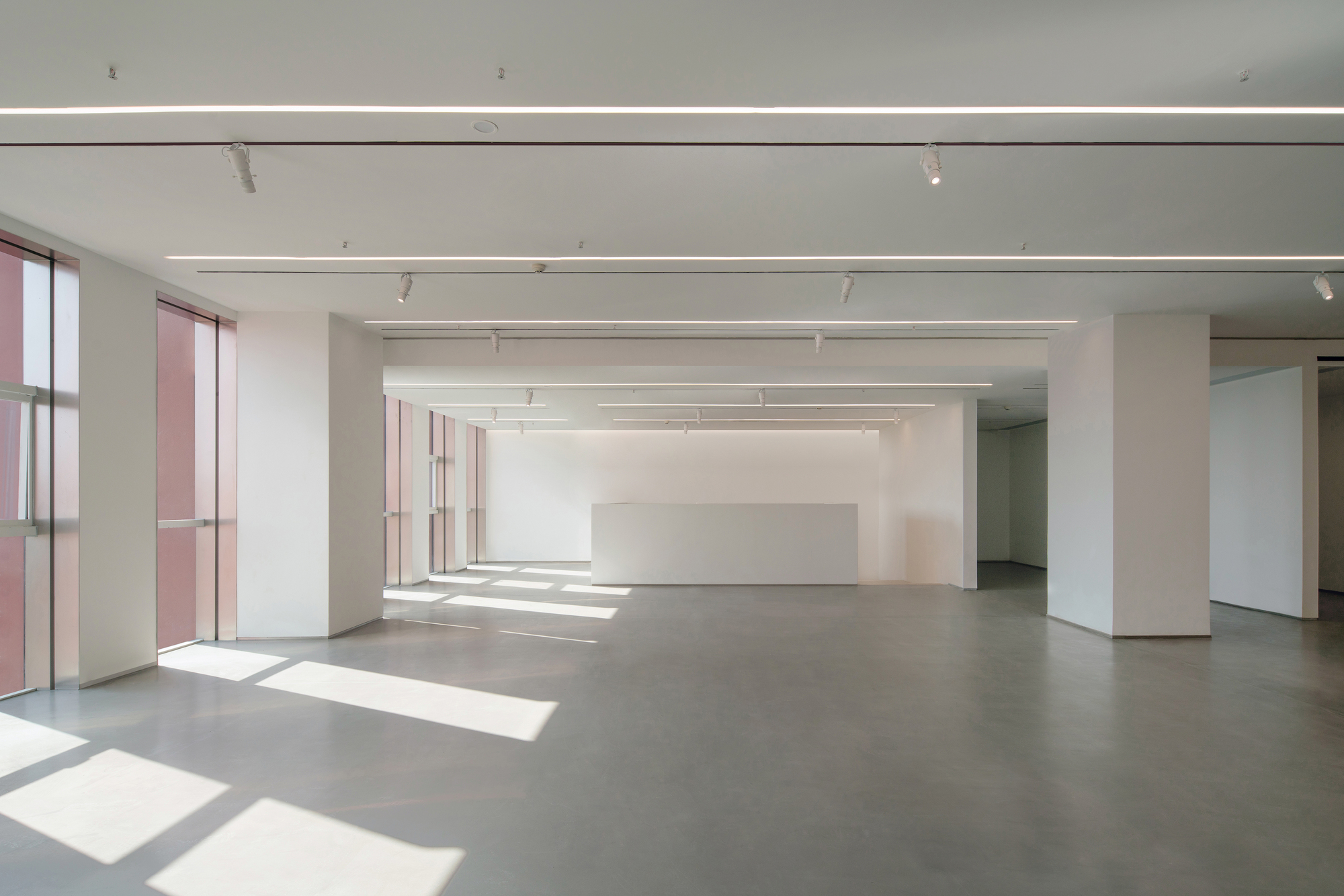 Open white exhibition floor with square columns, track lights and sunlight from pink-tinted windows falling across the concrete