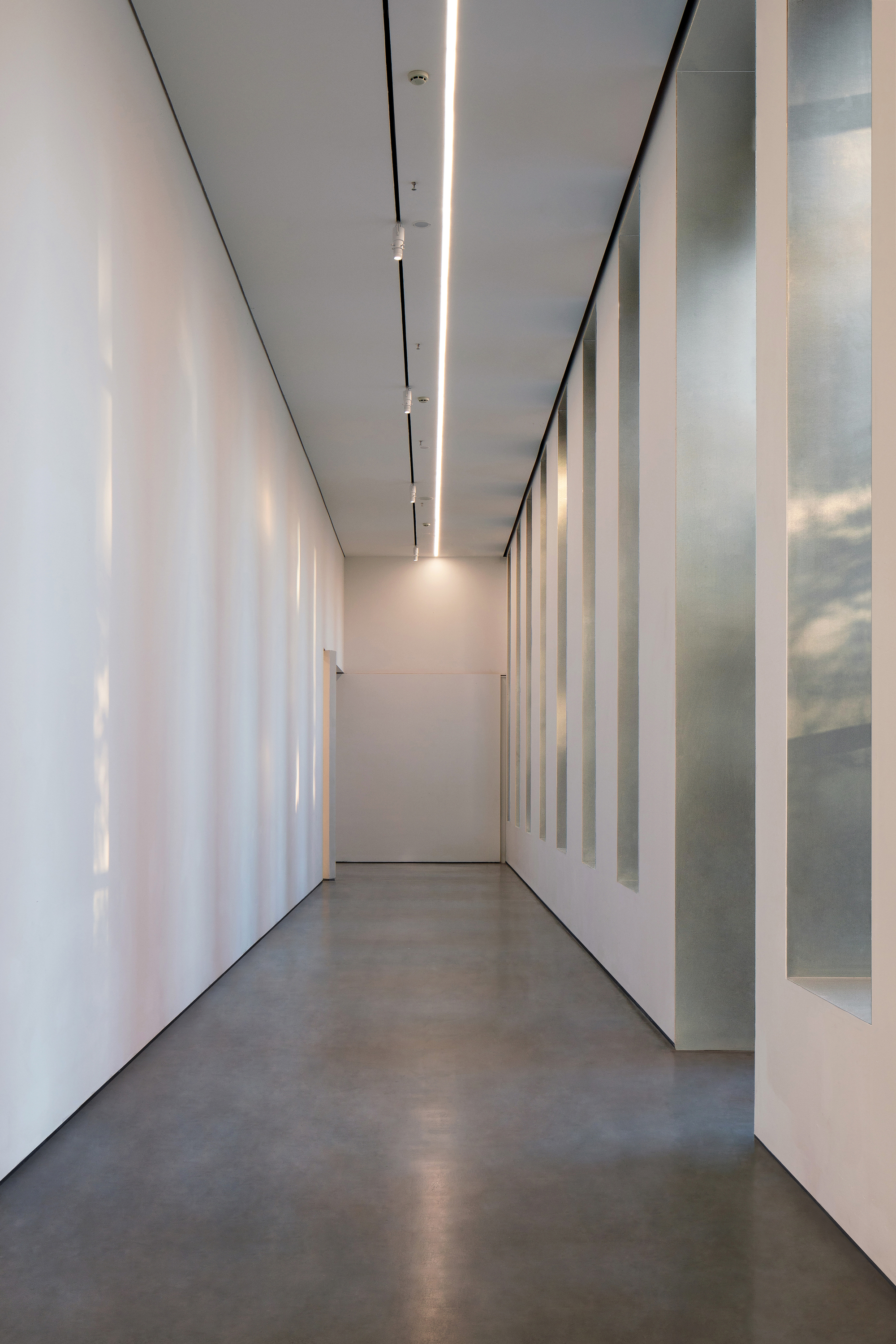 Narrow corridor with a blank white wall on one side and tall slot windows on the other, a linear light overhead