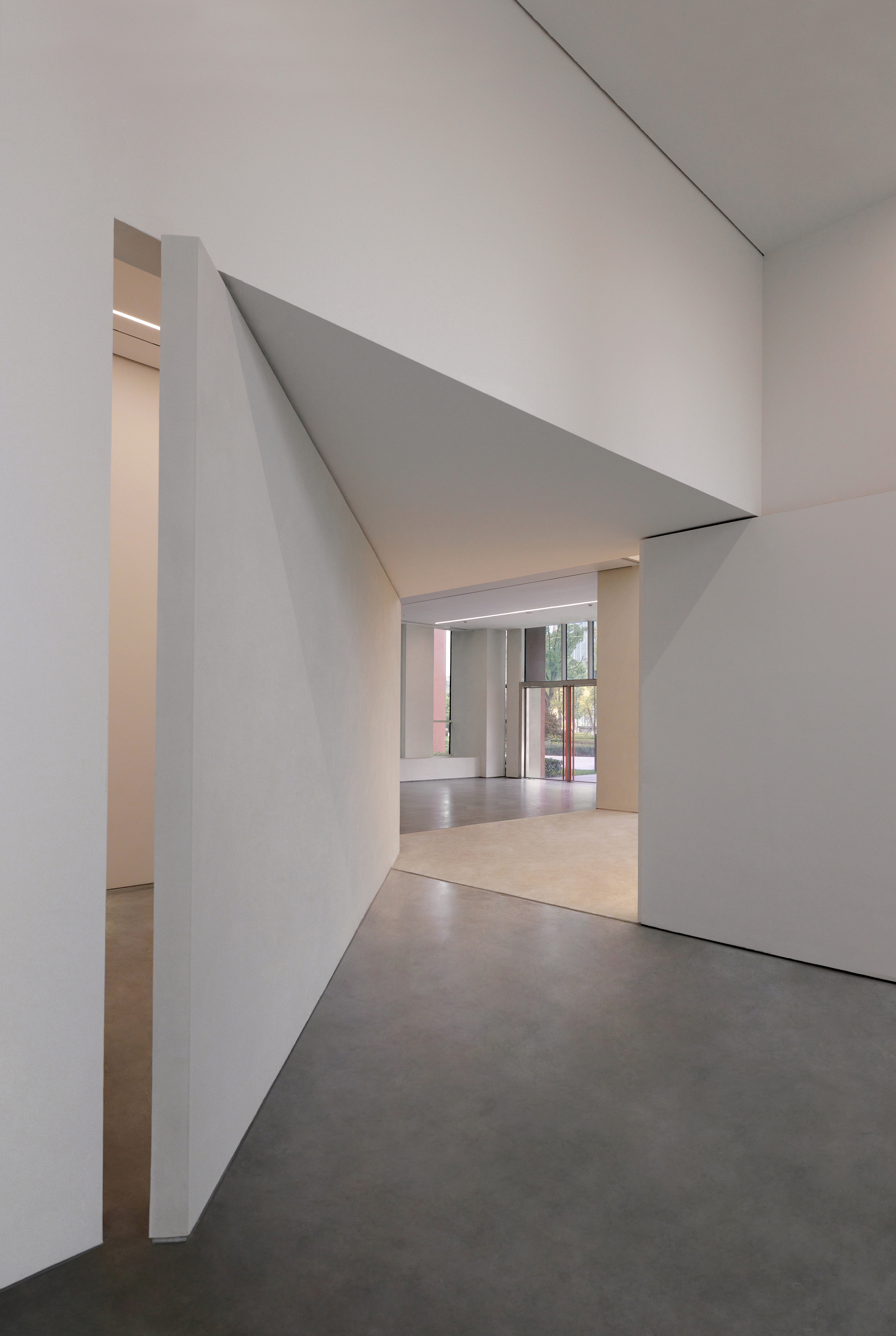 Angled white wall planes framing an opening toward a sunlit lobby with a pale stone floor patch and red-framed glazing