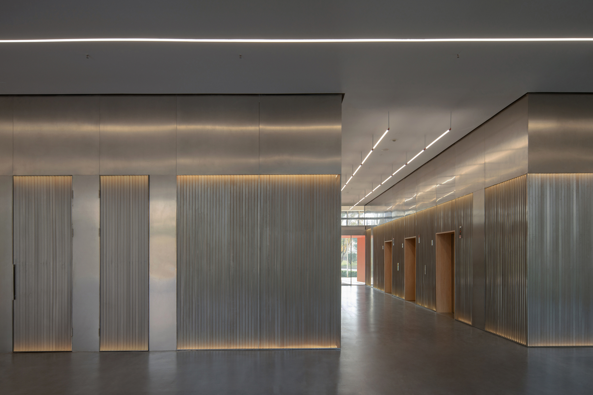 Corridor of fluted metal wall panels with warm backlit bases and timber-lined lift doors, ending at a glazed opening