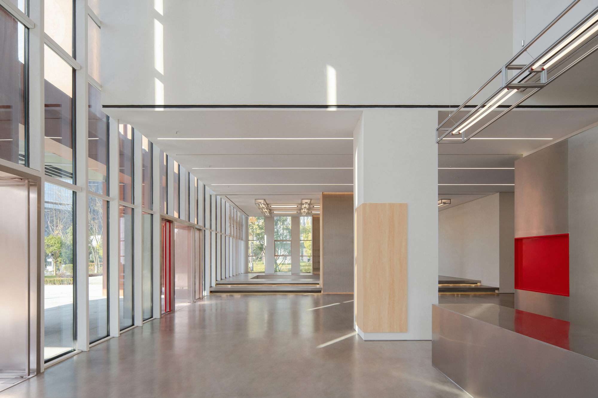 Double-height lobby with floor-to-ceiling glazing, a timber-clad column, a red panel and a steel counter in afternoon sun