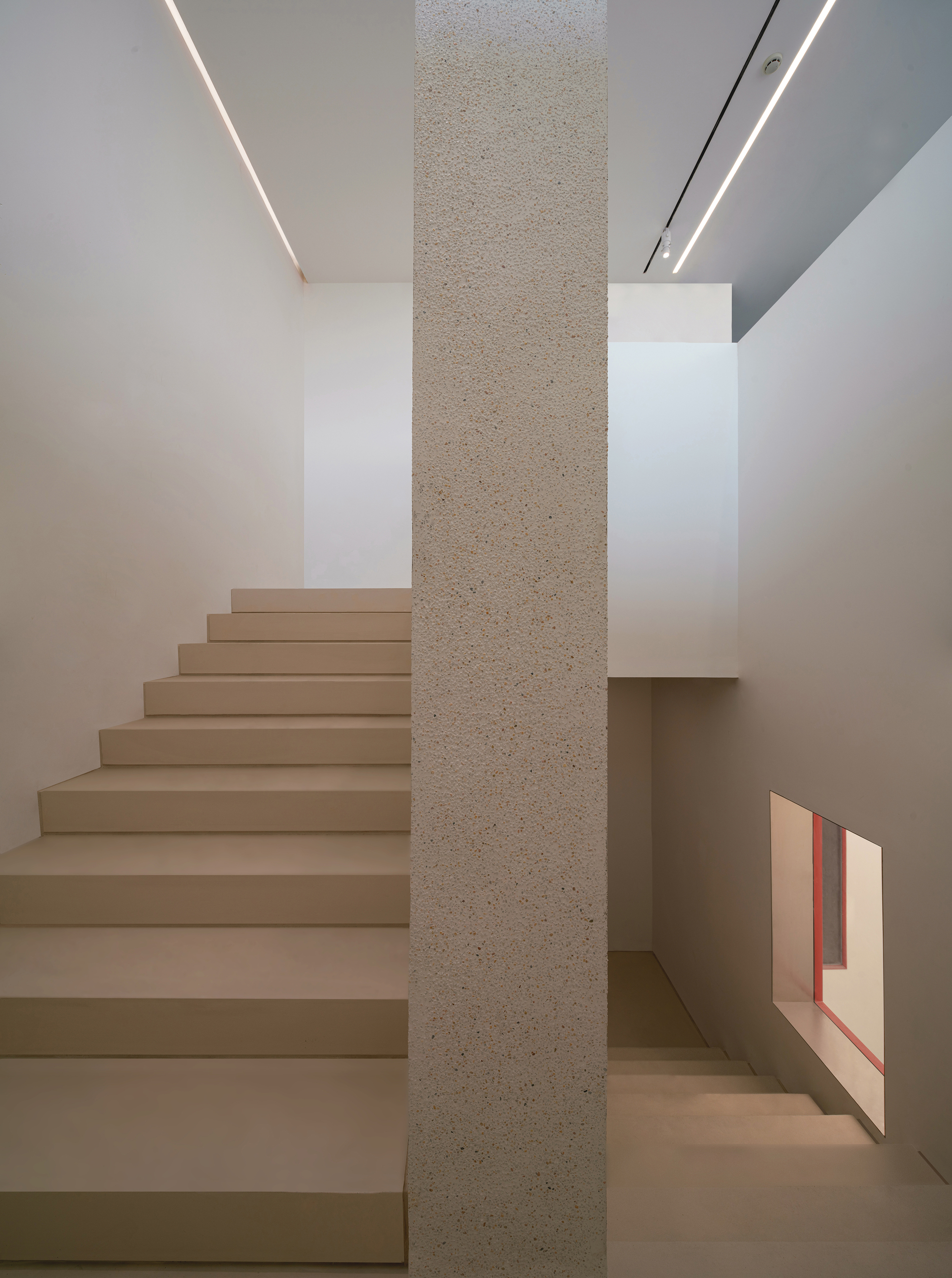 Straight stair of pale stone treads beside a speckled concrete column, with linear ceiling lights and a small red-framed window opening