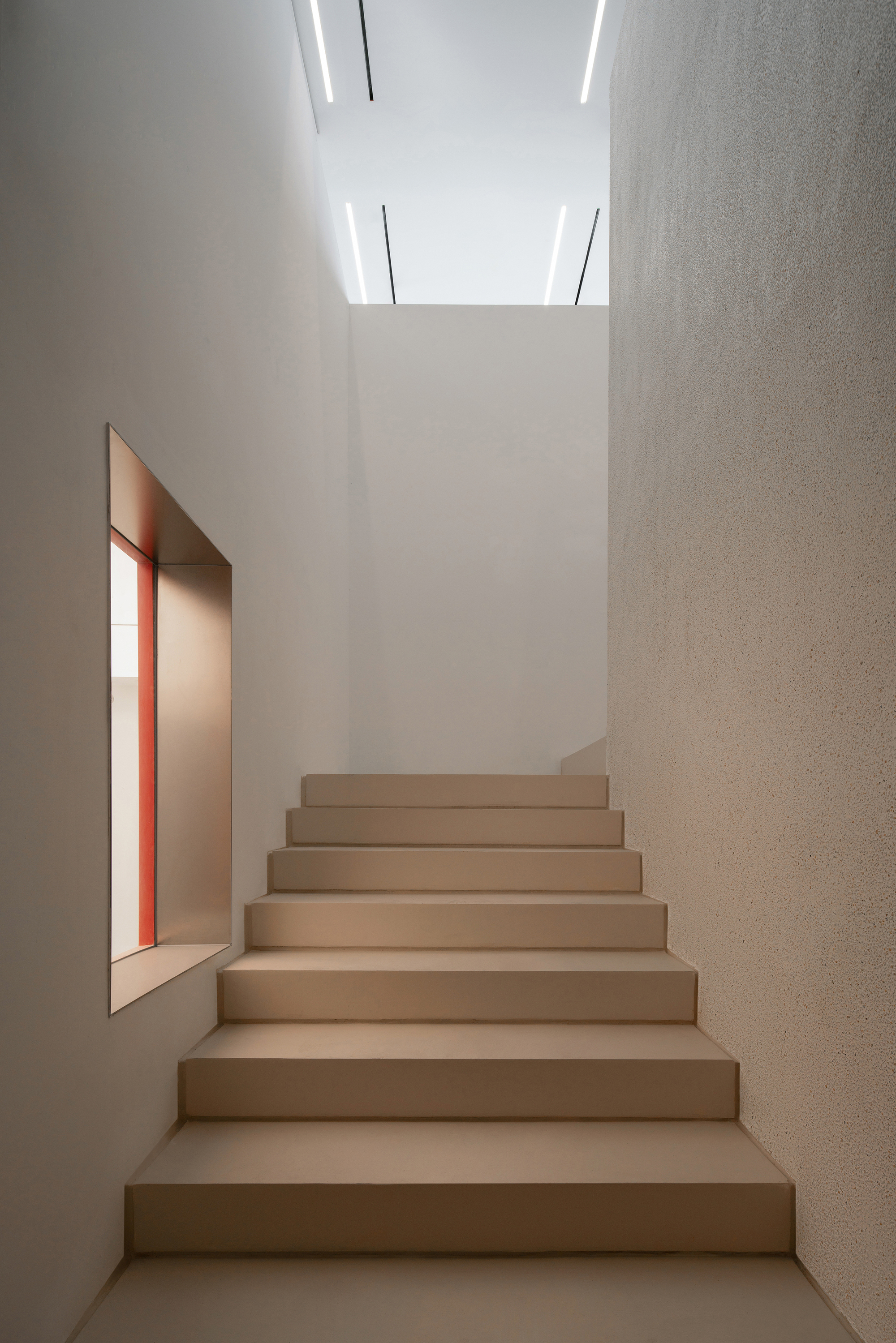 Narrow stair of beige treads rising between plaster walls, with a red-lined window recess and linear ceiling lights above