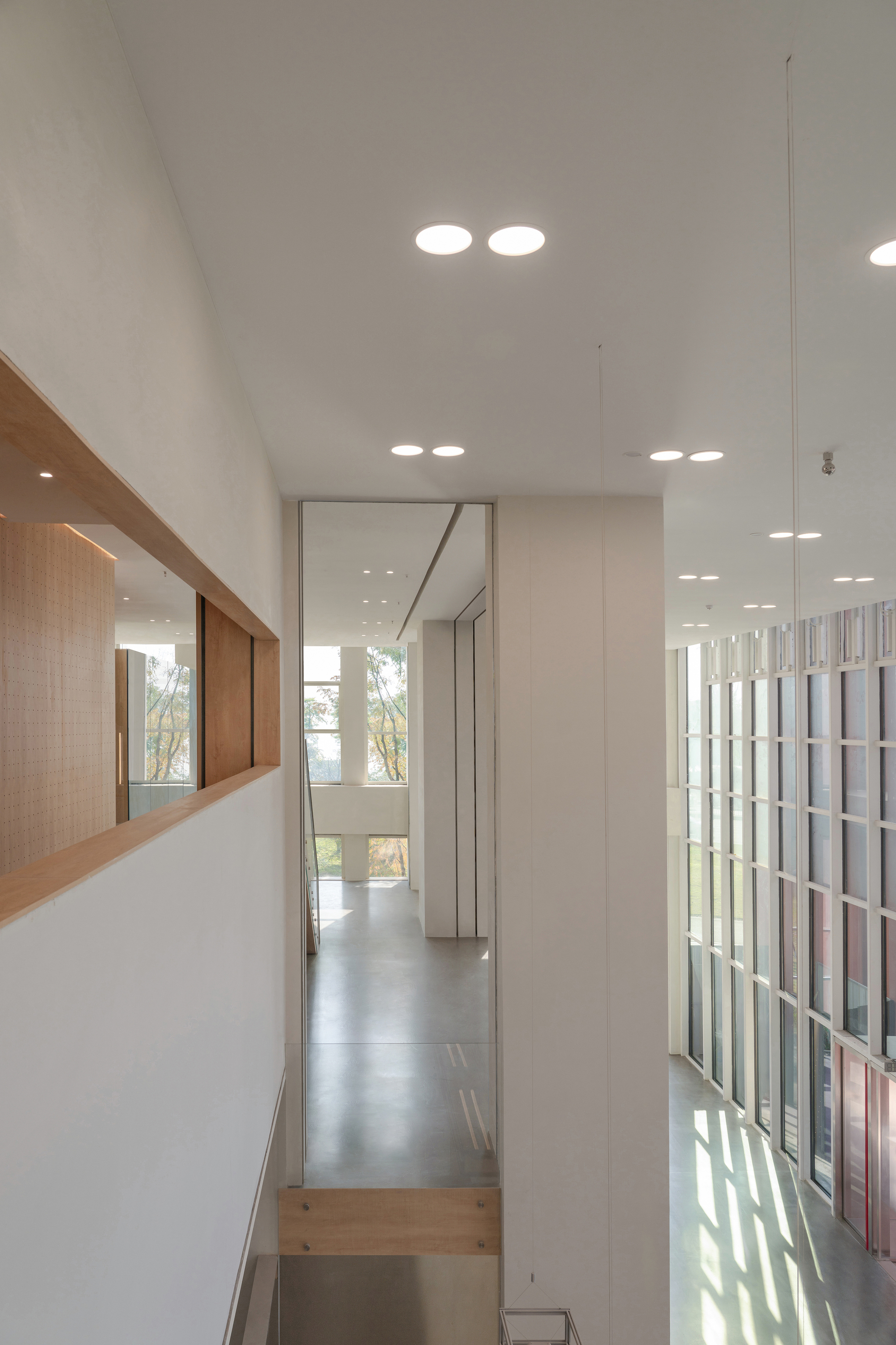 View from an upper level through a timber-framed wall opening toward a glazed facade and sunlit concrete floor