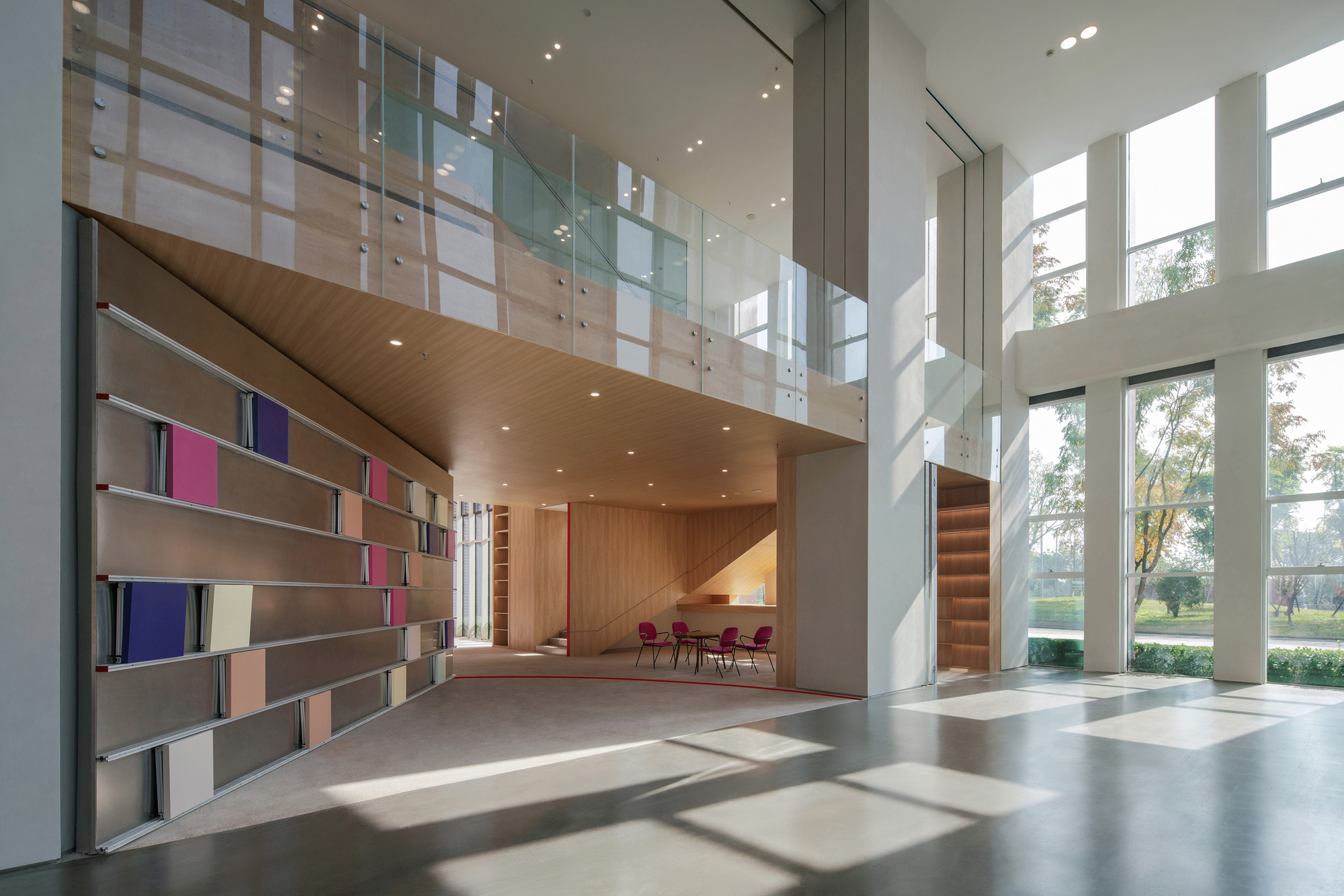 Double-height lobby with a shelf wall of colored panels, a glass balustrade above and autumn trees outside tall windows