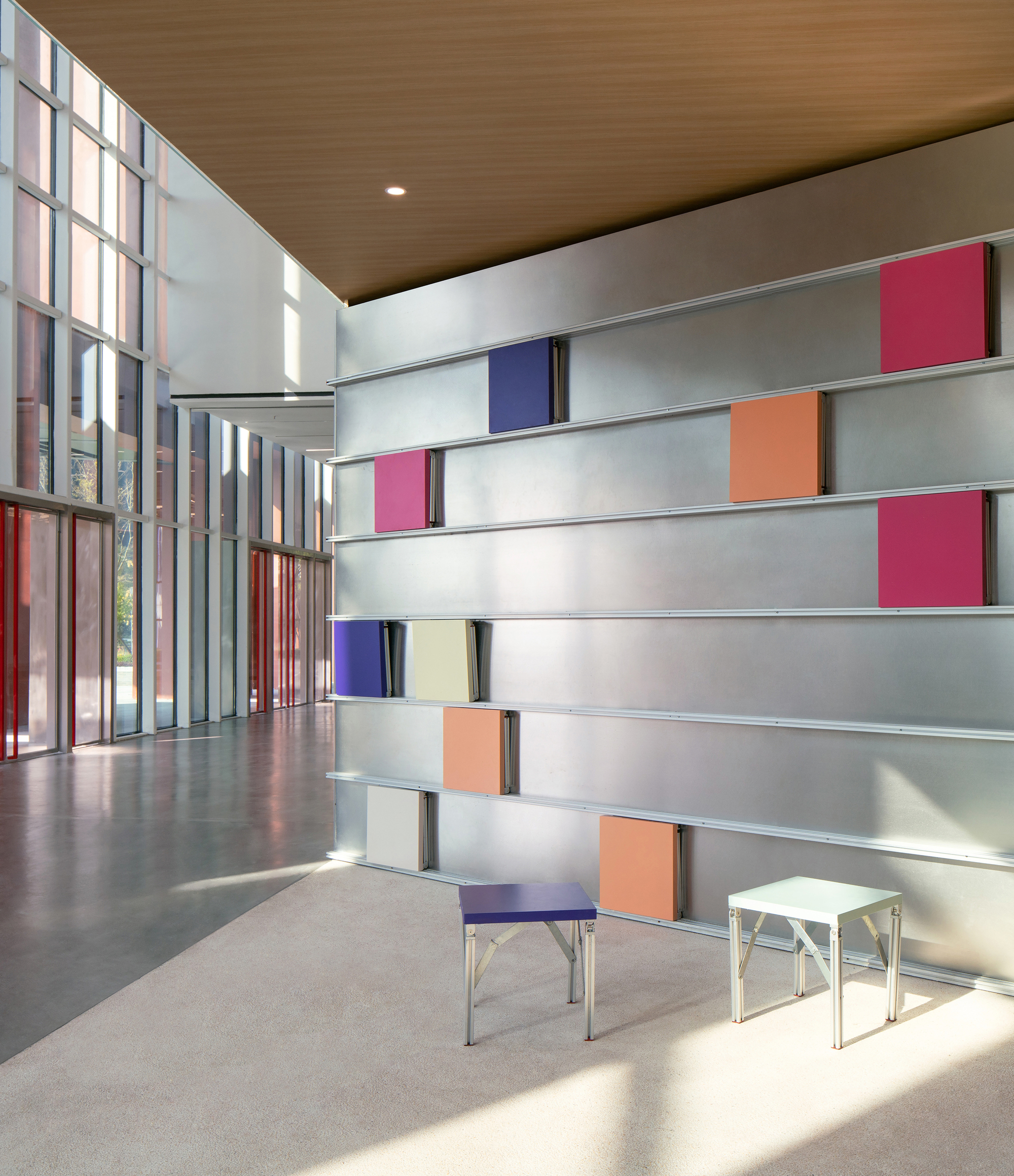 Metal wall of horizontal rails holding pink, blue and orange panels, with two small stools in sunlight on the carpet