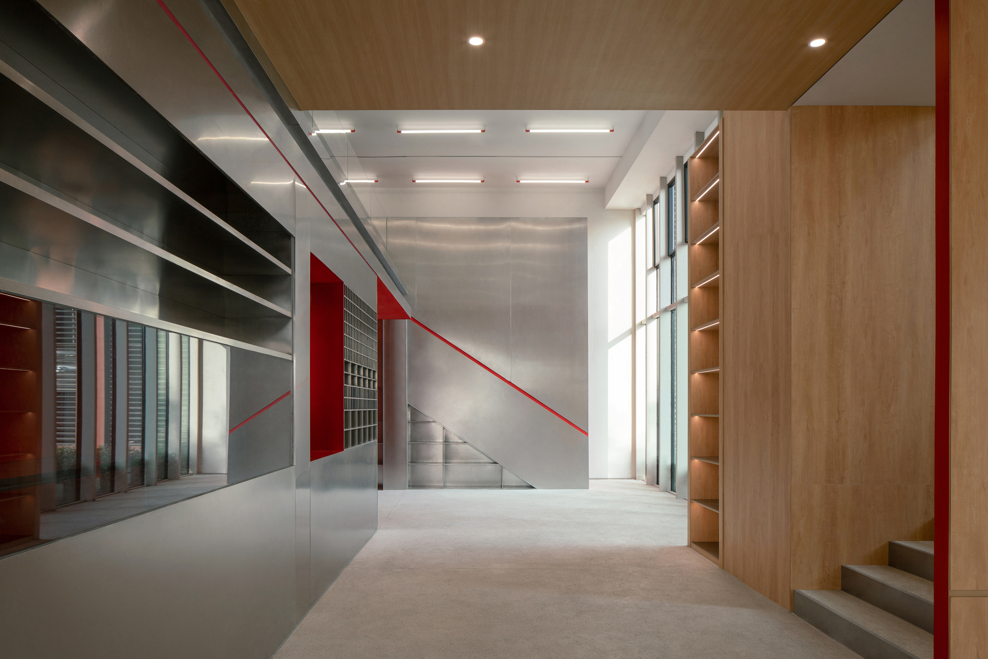 Hallway of brushed steel panels and timber shelving with a red stripe tracing the stair, lit by daylight from a tall window slot