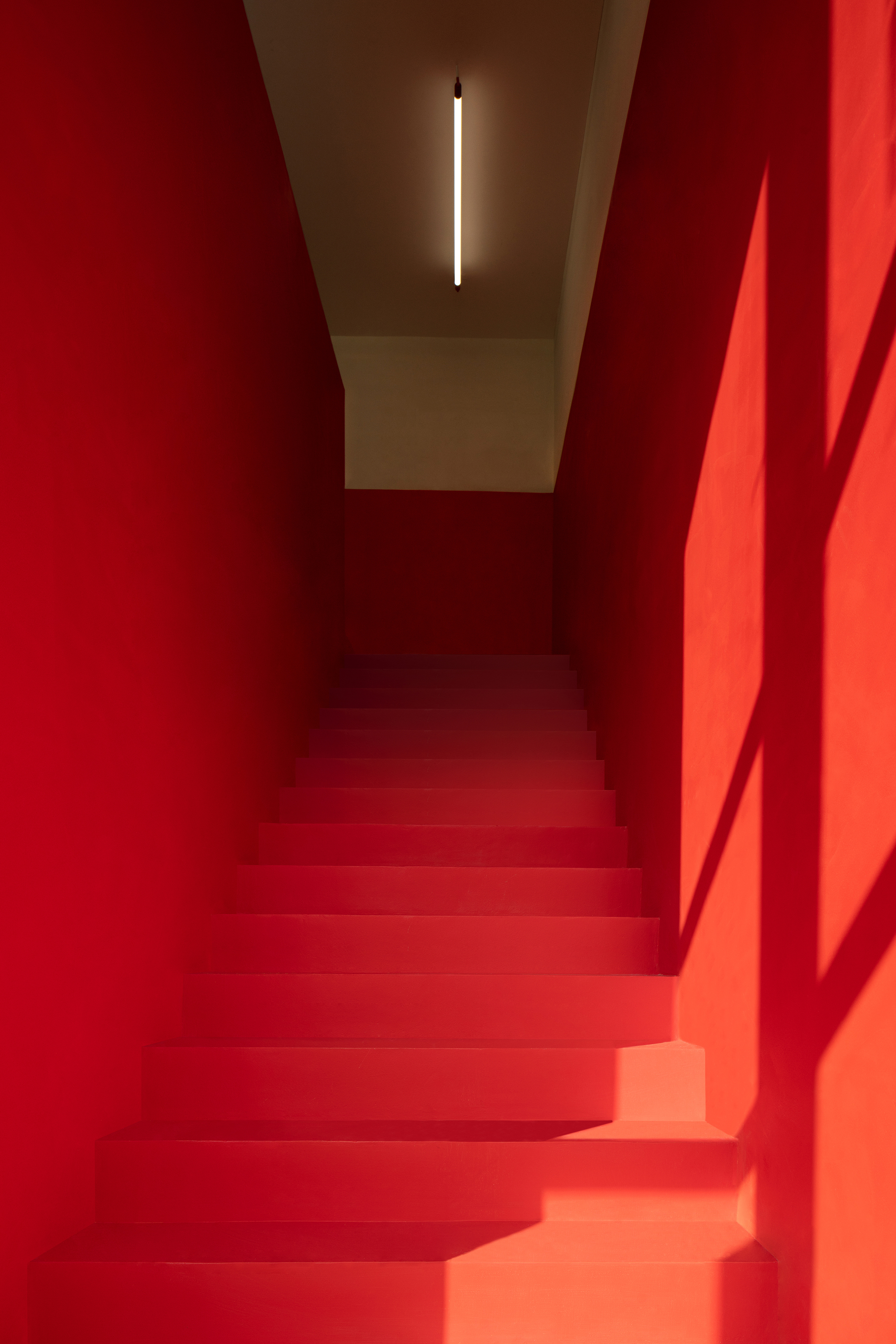 Narrow red staircase rising toward a single linear light fixture, with window shadows cast across the right wall in low sun