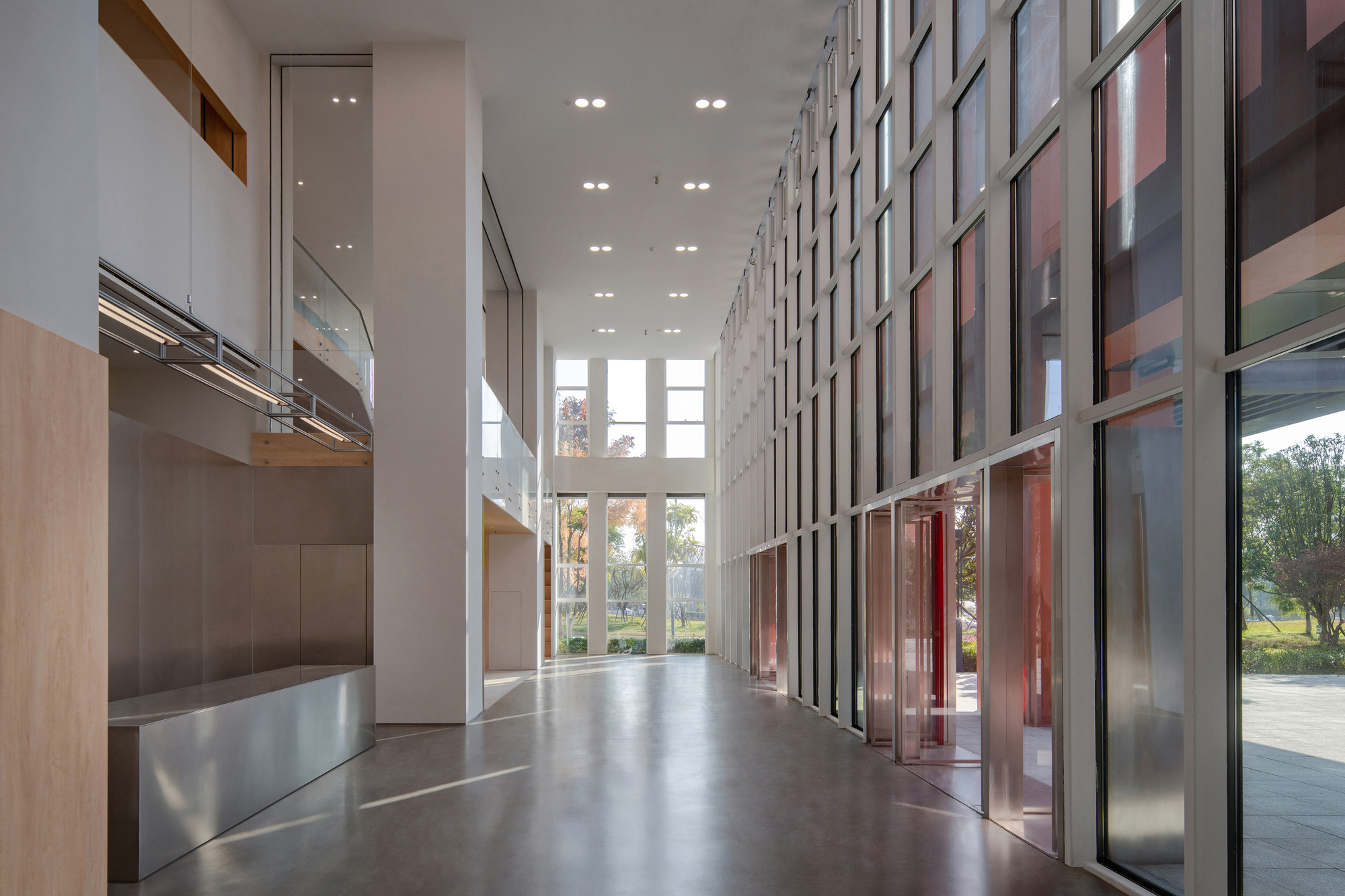 Double-height lobby with a full-height glazed wall, steel bench and white columns, trees visible outside in soft daylight