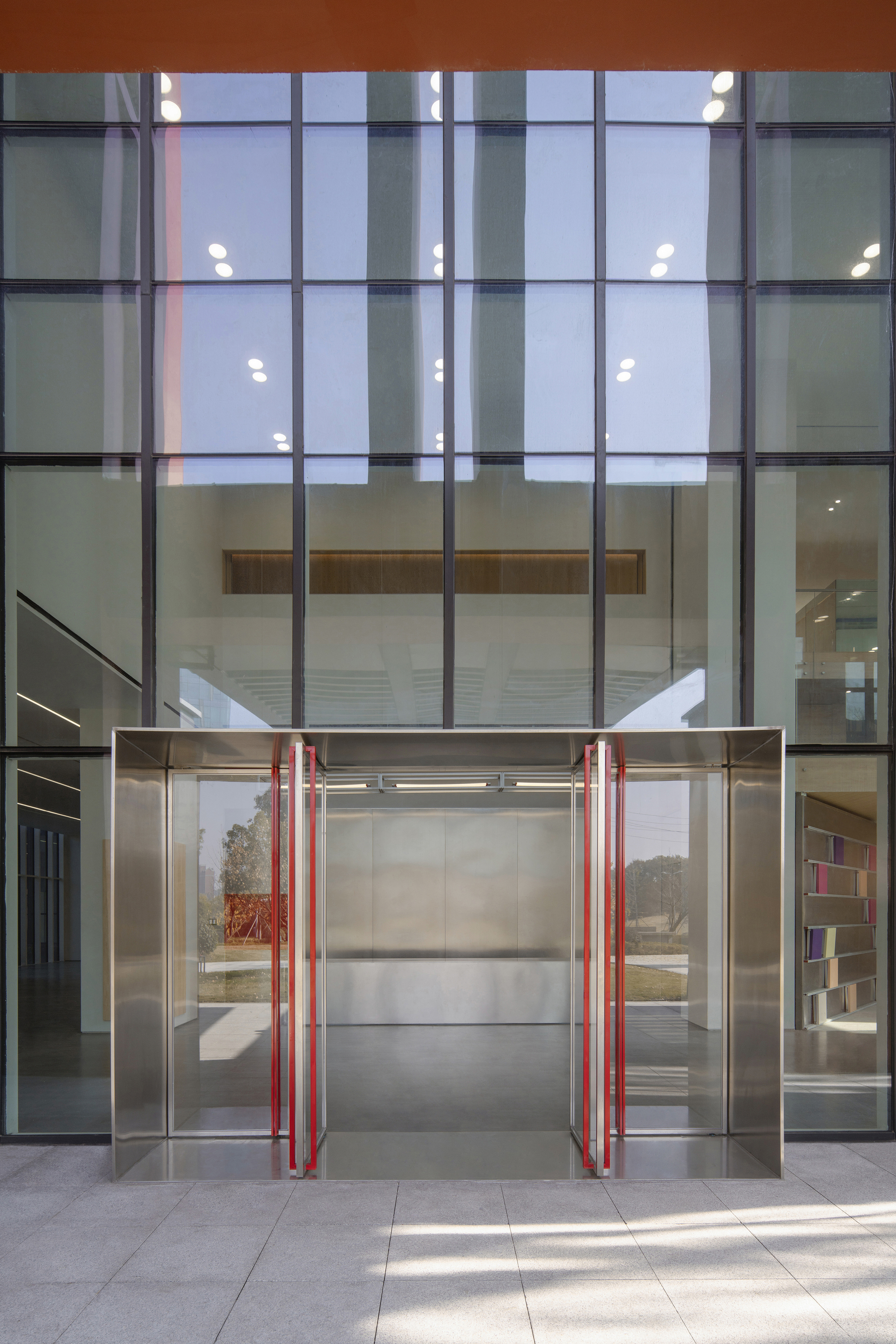 Stainless steel entrance vestibule with red door handles set into a tall glass curtain wall above a paved forecourt