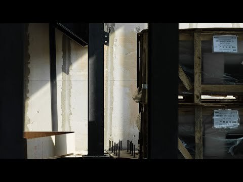 Construction site view of black steel columns and anchor bolts beside wrapped crates, with a strip of sunlight on the concrete wall