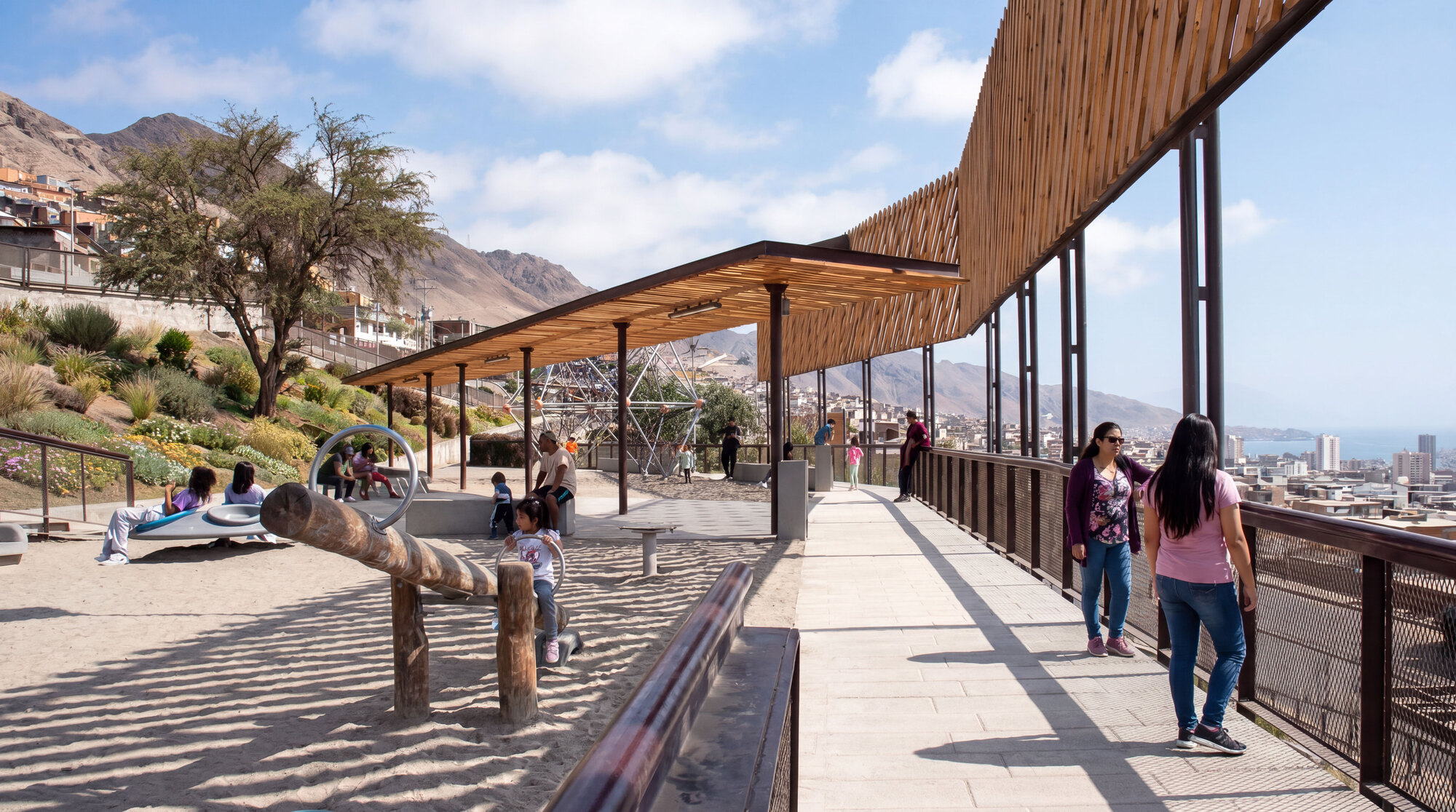 Timber-roofed walkway beside a sand play area with a log seesaw, as visitors stroll above a hillside city in bright sun