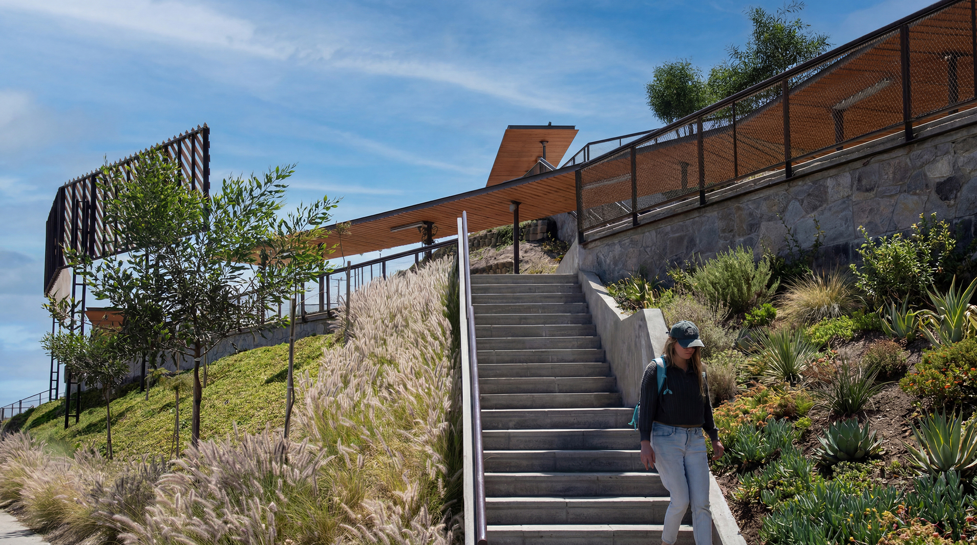 Concrete stairway climbing between a stone retaining wall and grasses toward a slatted timber canopy, with a walker in a cap under blue sky