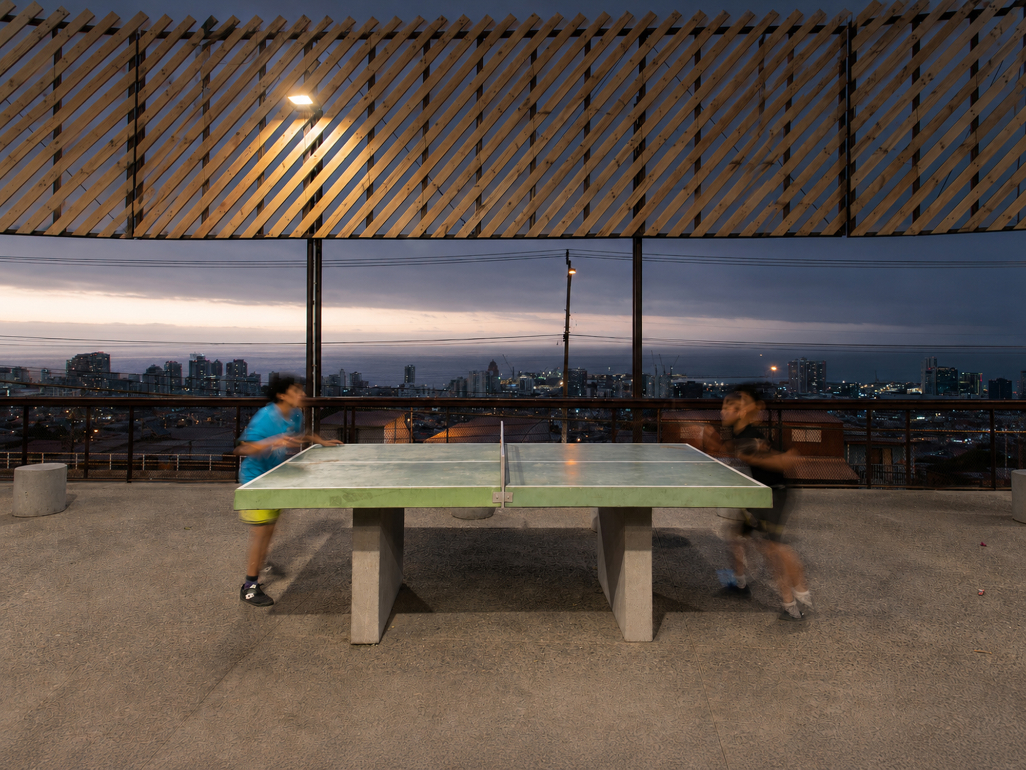 Two blurred children playing at a concrete ping-pong table beneath a slatted timber screen at dusk, with city lights behind