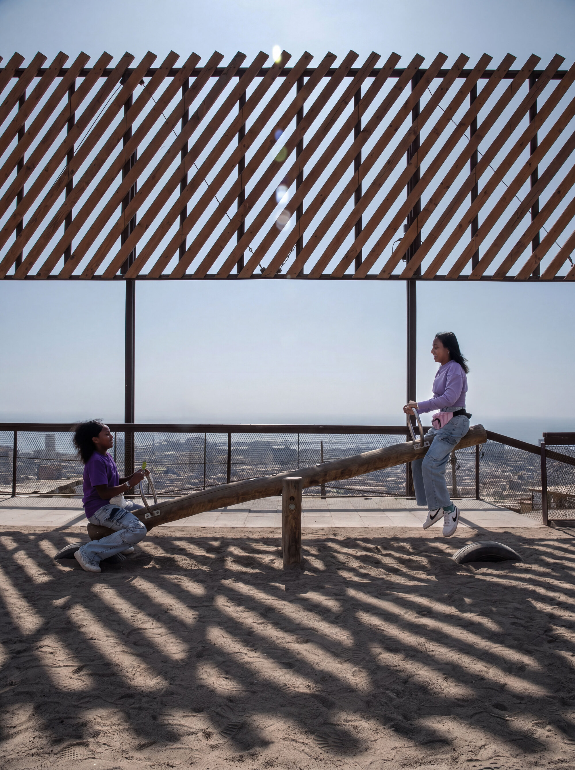 Two girls on a log seesaw over sand, with diagonal timber slat shadows falling across the ground in hazy midday sun