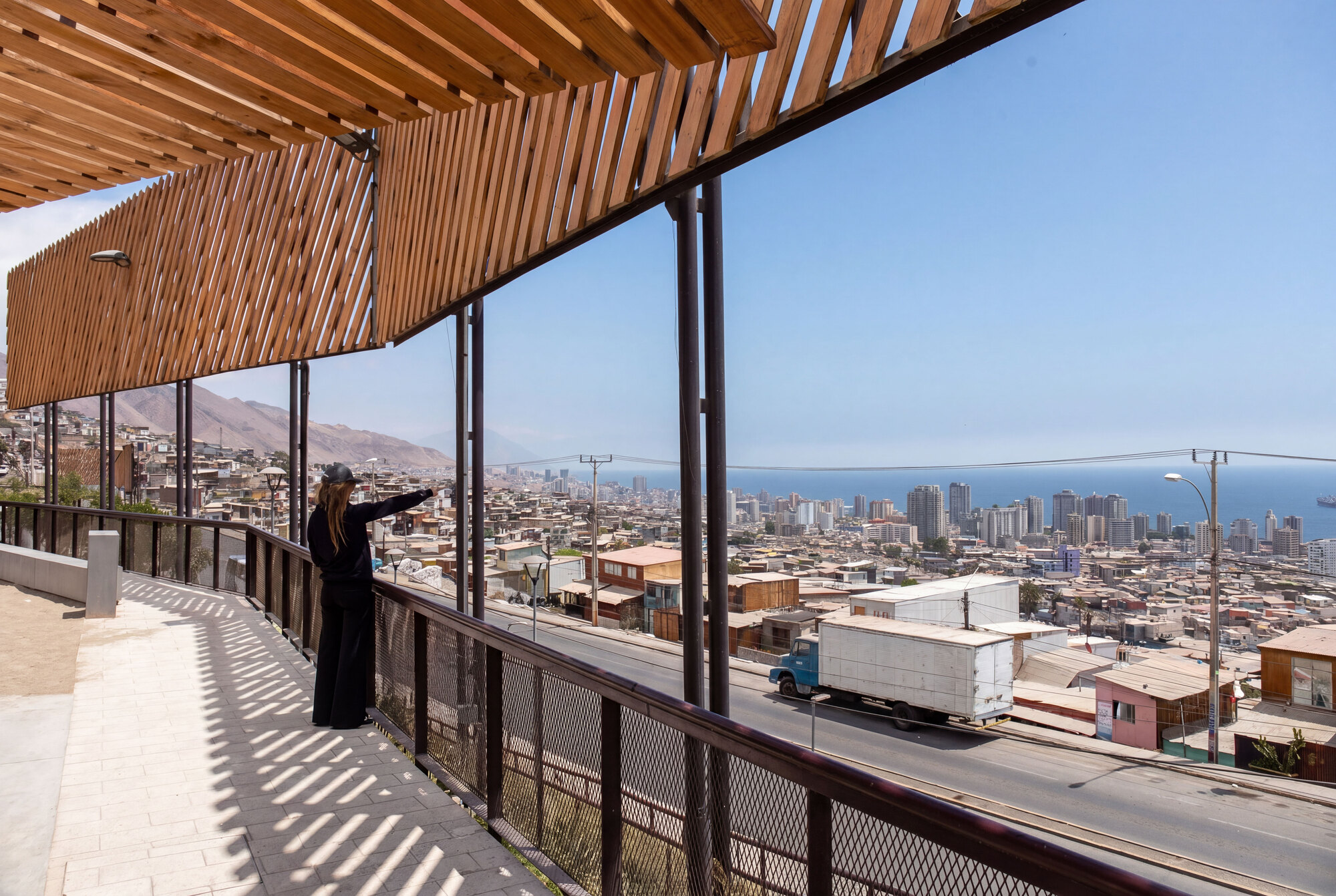 Viewing terrace under a slatted timber canopy, a woman at the steel railing facing a hillside city and distant ocean in bright sun