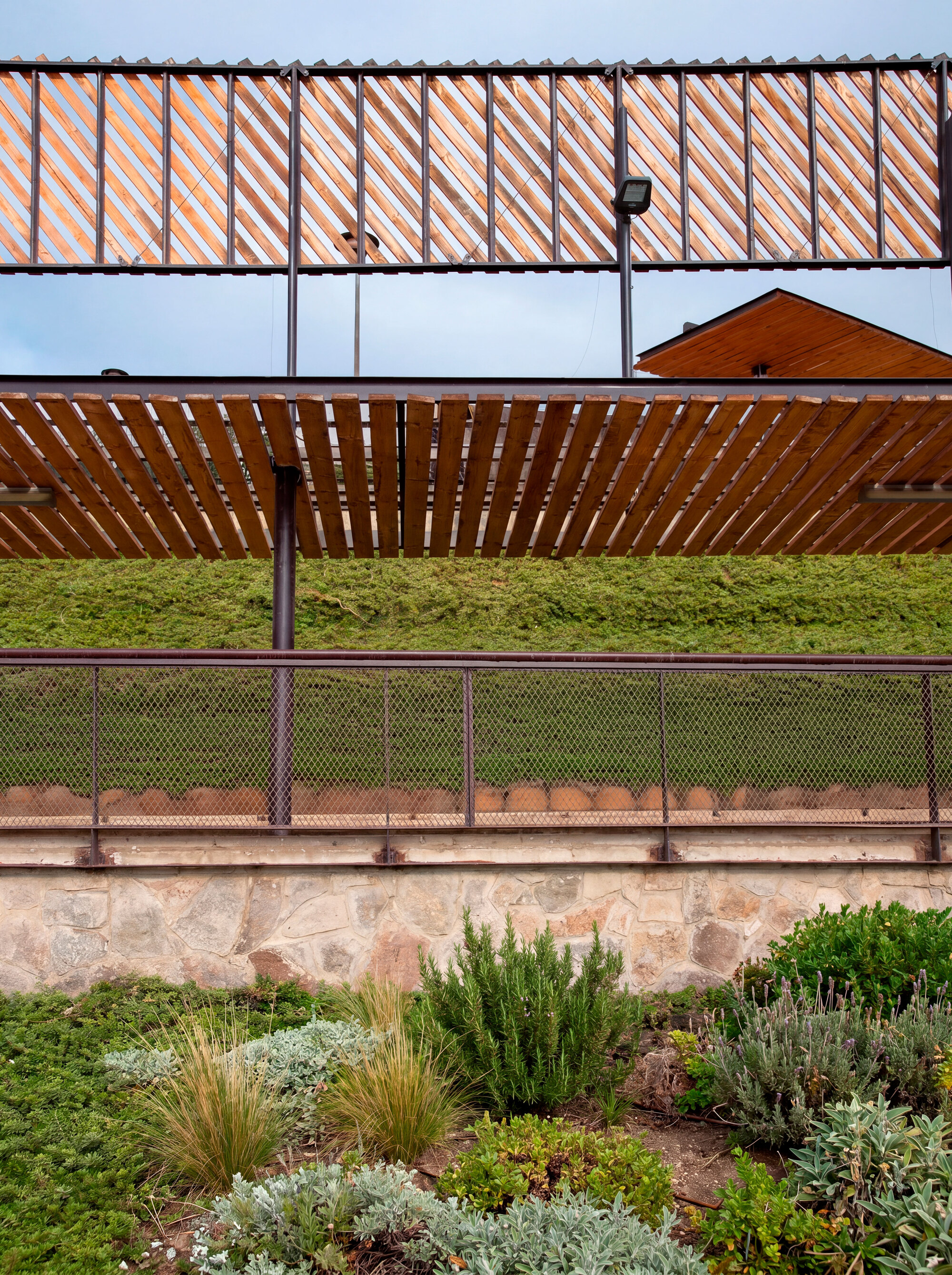 Close-up of layered slat canopies and mesh railing over a stone wall, with rosemary and grasses in the foreground under a cloudy sky