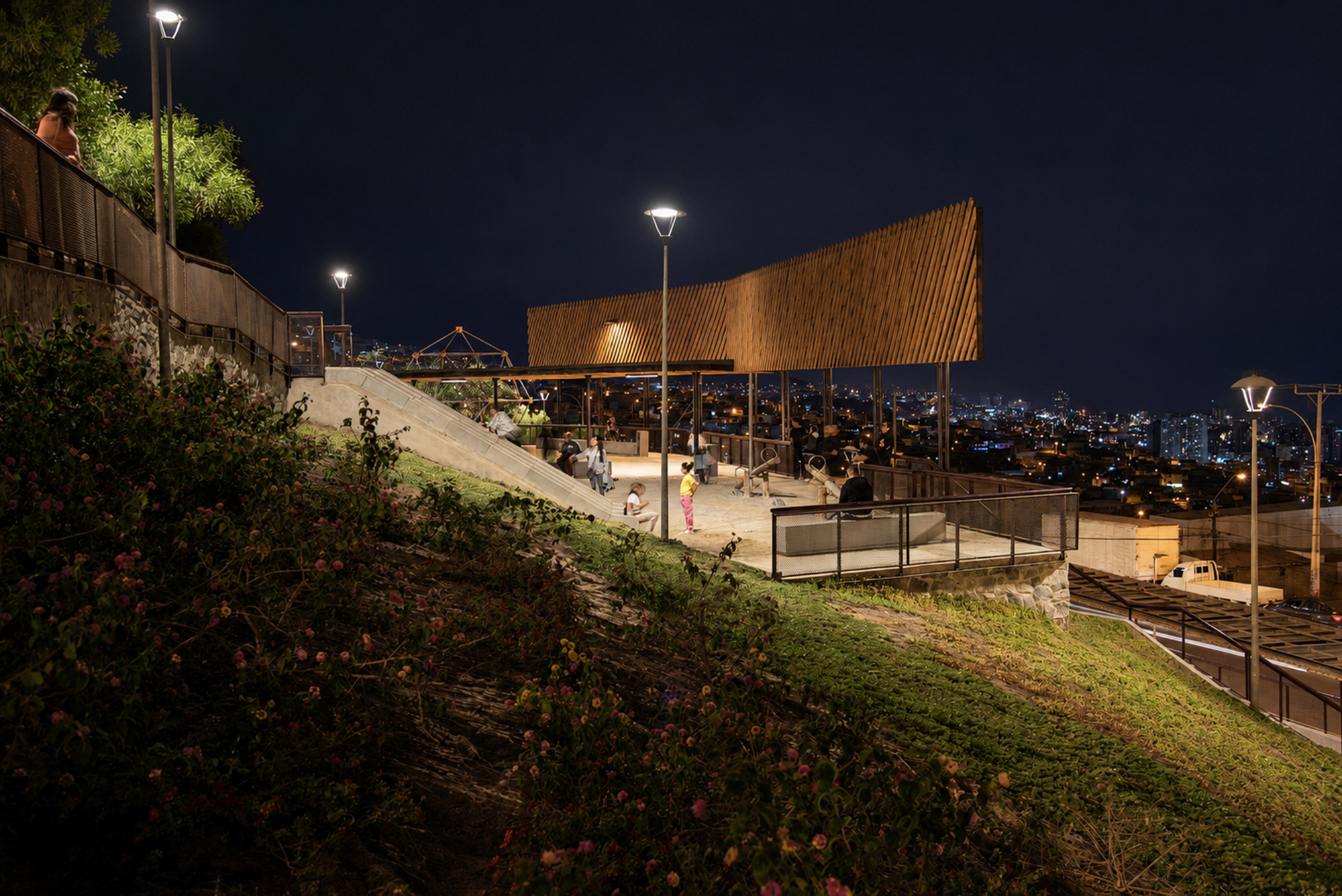 Dusk view of a slatted timber screen over a hilltop plaza, with a grassy embankment and city lights spread below