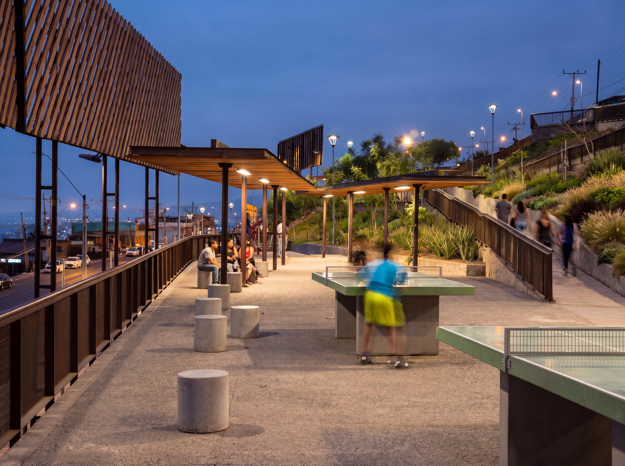Concrete table tennis tables and round stools under timber-slatted canopies on a hillside promenade at dusk, a blurred child mid-game
