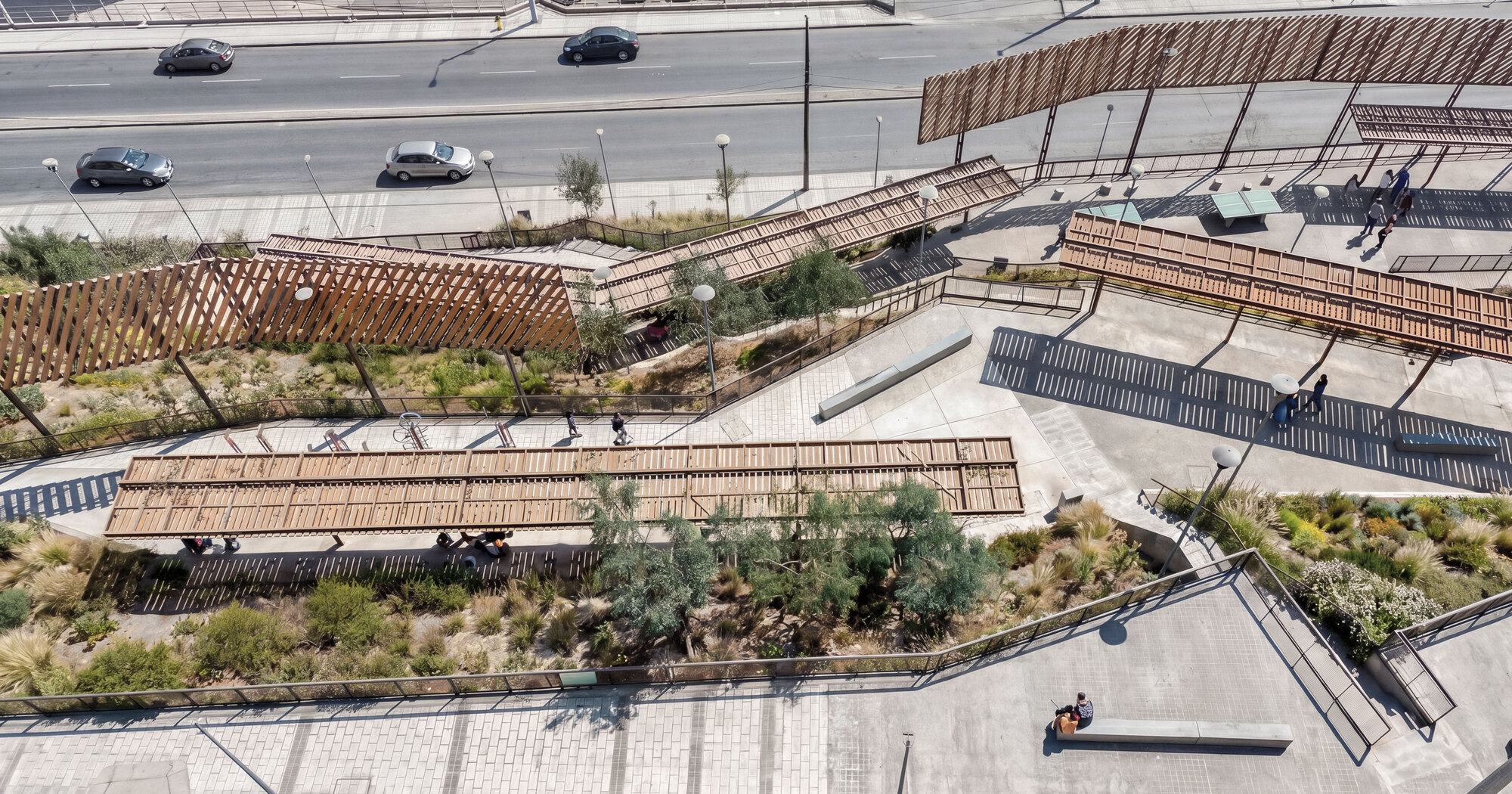 Aerial view of slatted timber shade roofs and planted slopes stepping down beside a busy road in daylight