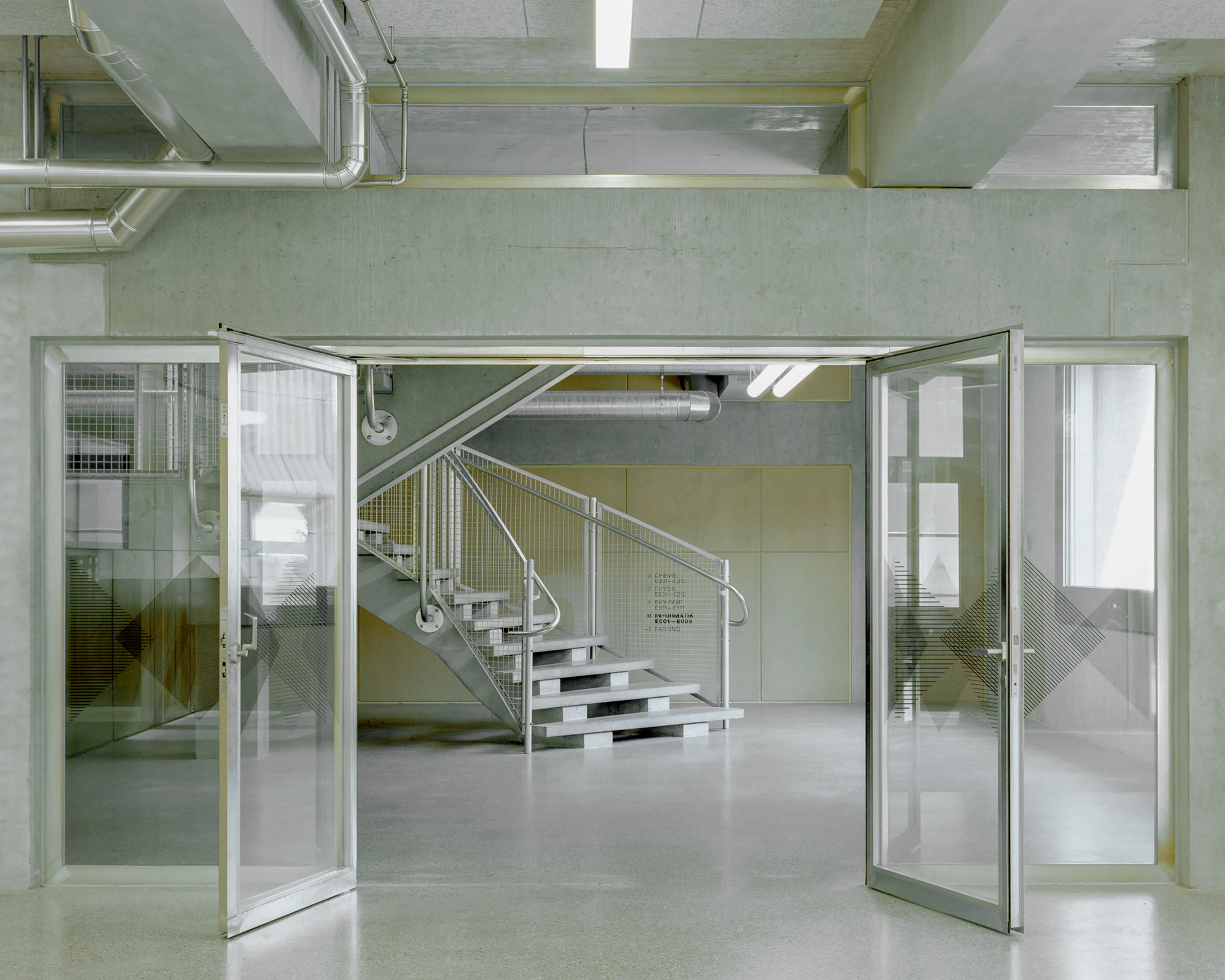 Open glass double doors framing a galvanized steel staircase beyond, with exposed ducts and concrete beams overhead under fluorescent tubes