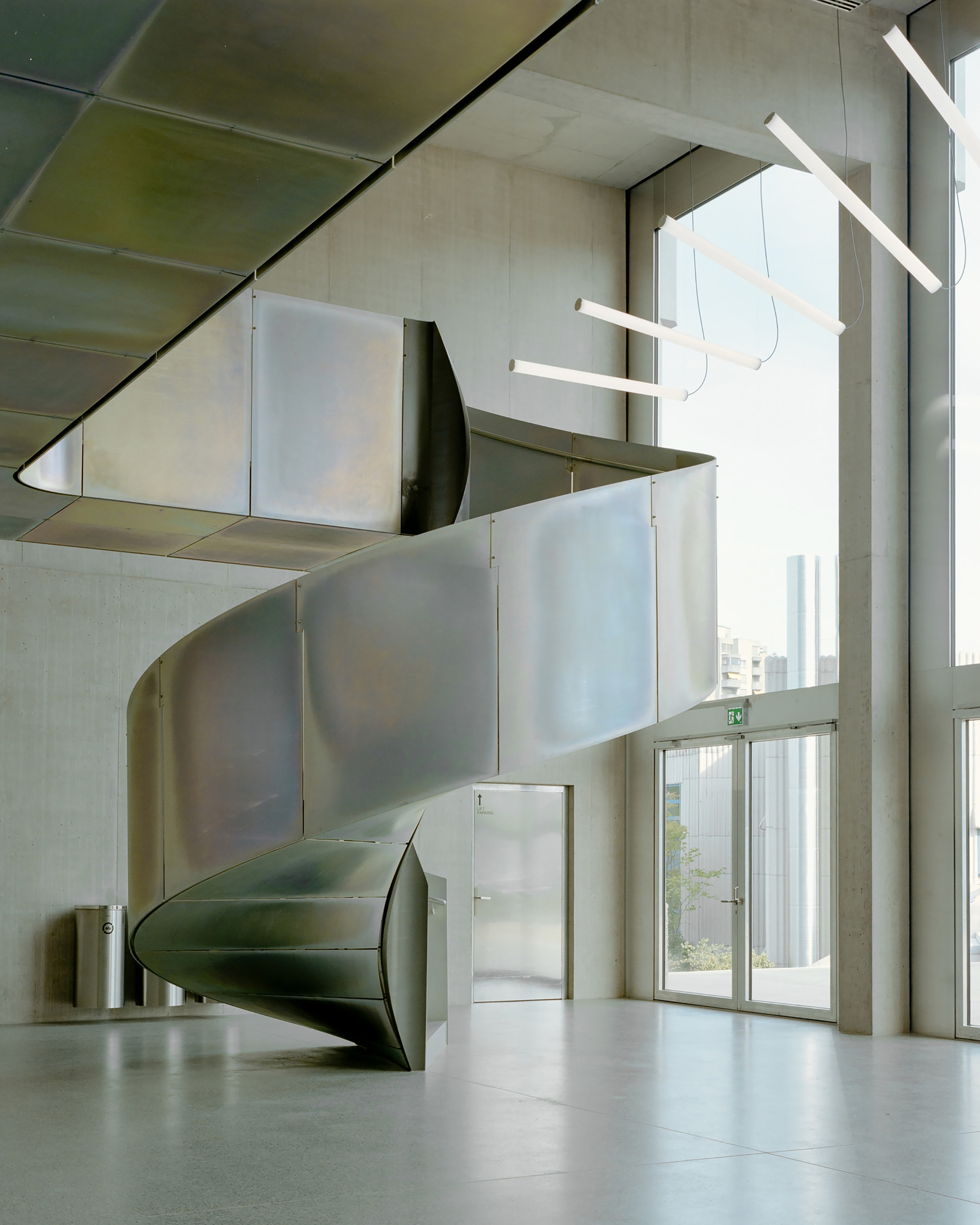 Spiral staircase clad in brushed steel panels rising through a tall concrete hall beside a glazed wall with city views