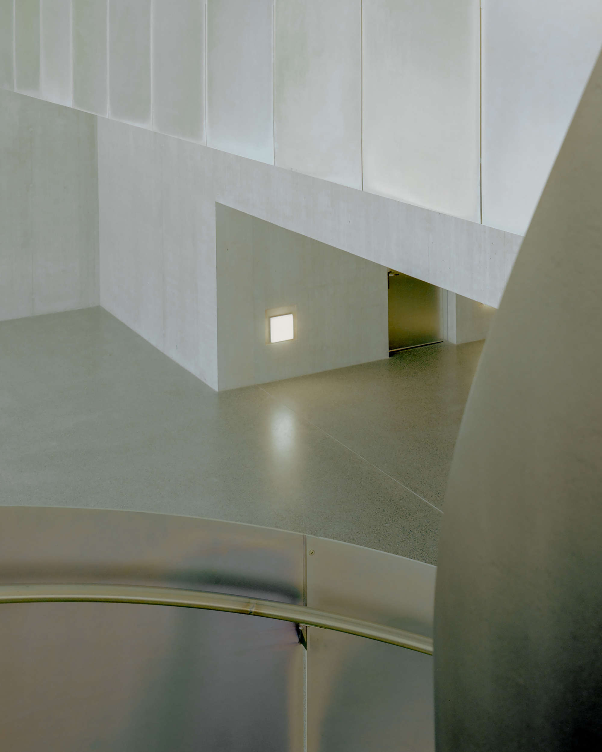 Detail of a concrete lobby with a lit wall light and a dark doorway, framed by the curved steel stair balustrade
