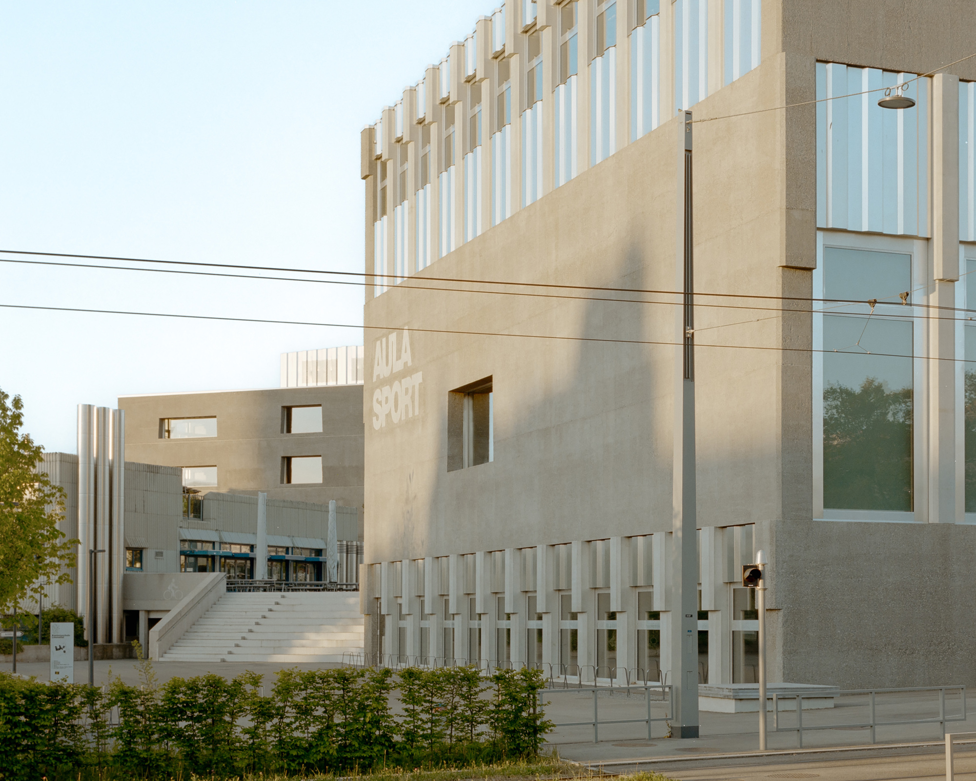 Street corner view of a concrete building with the lettering AULA SPORT, ribbed upper facade and wide steps, beside overhead tram wires