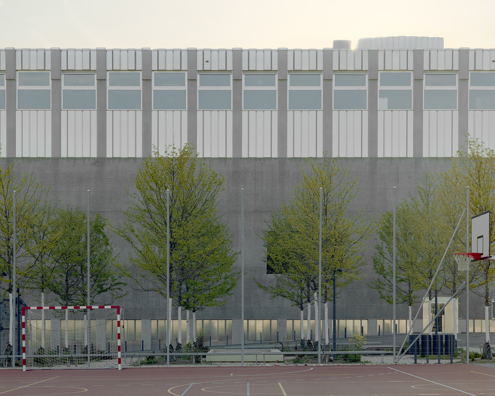Long concrete wall below a corrugated panel and window band, seen past young trees and a basketball court at dusk
