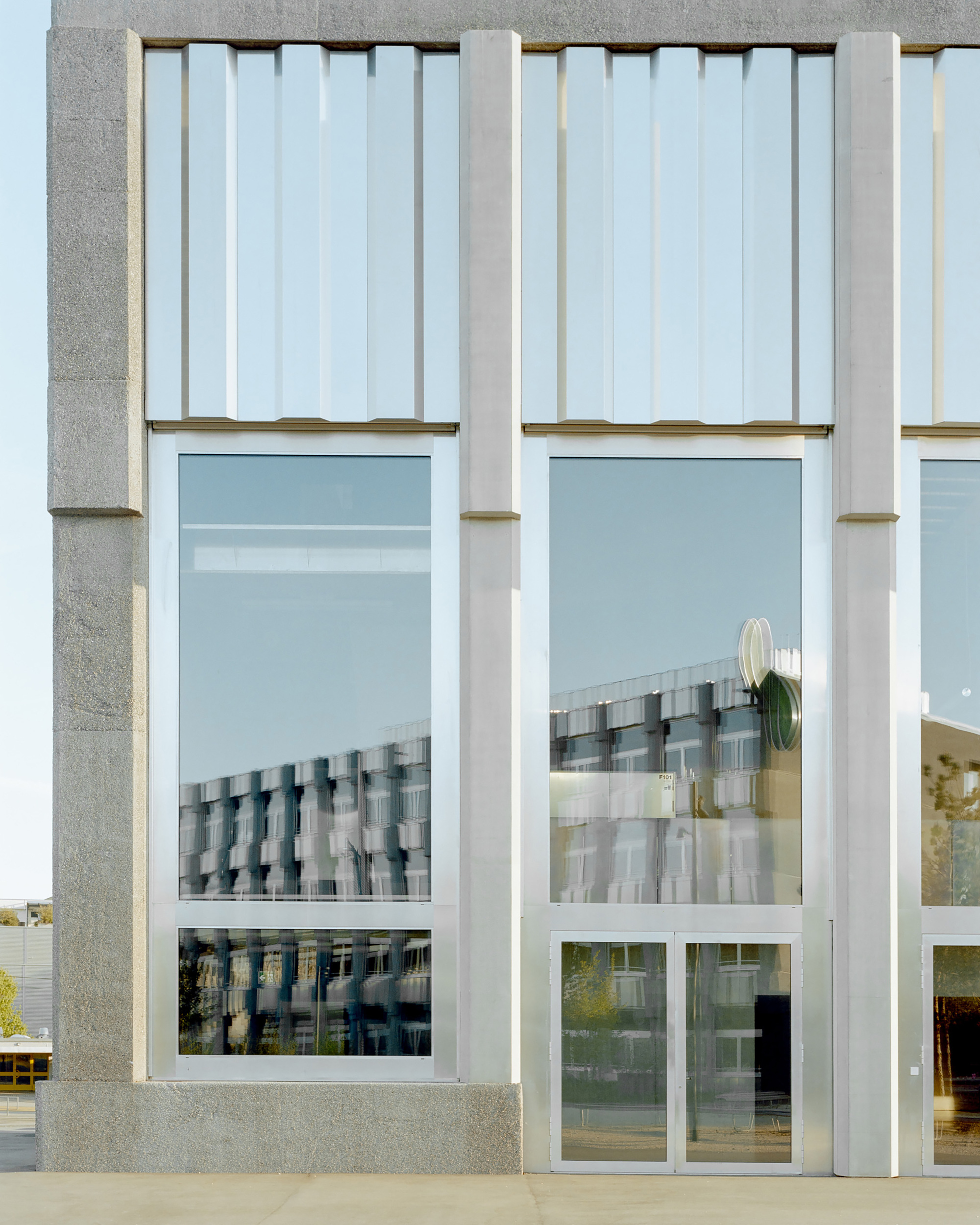Facade bay with concrete pilasters and tall glazing reflecting a neighbouring building under a clear blue sky