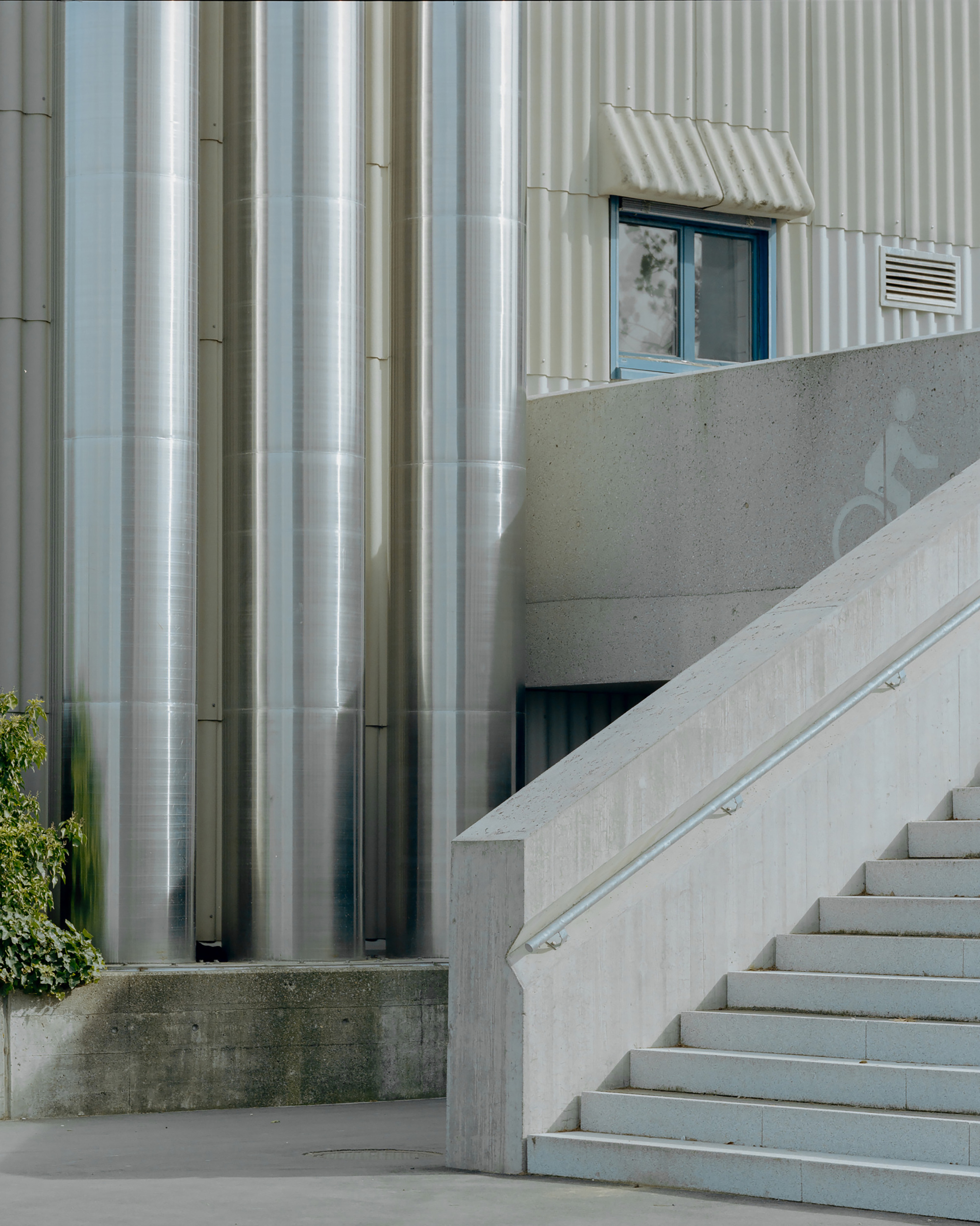 Polished metal cylindrical columns beside a concrete stair with handrail and a corrugated wall, in soft daylight