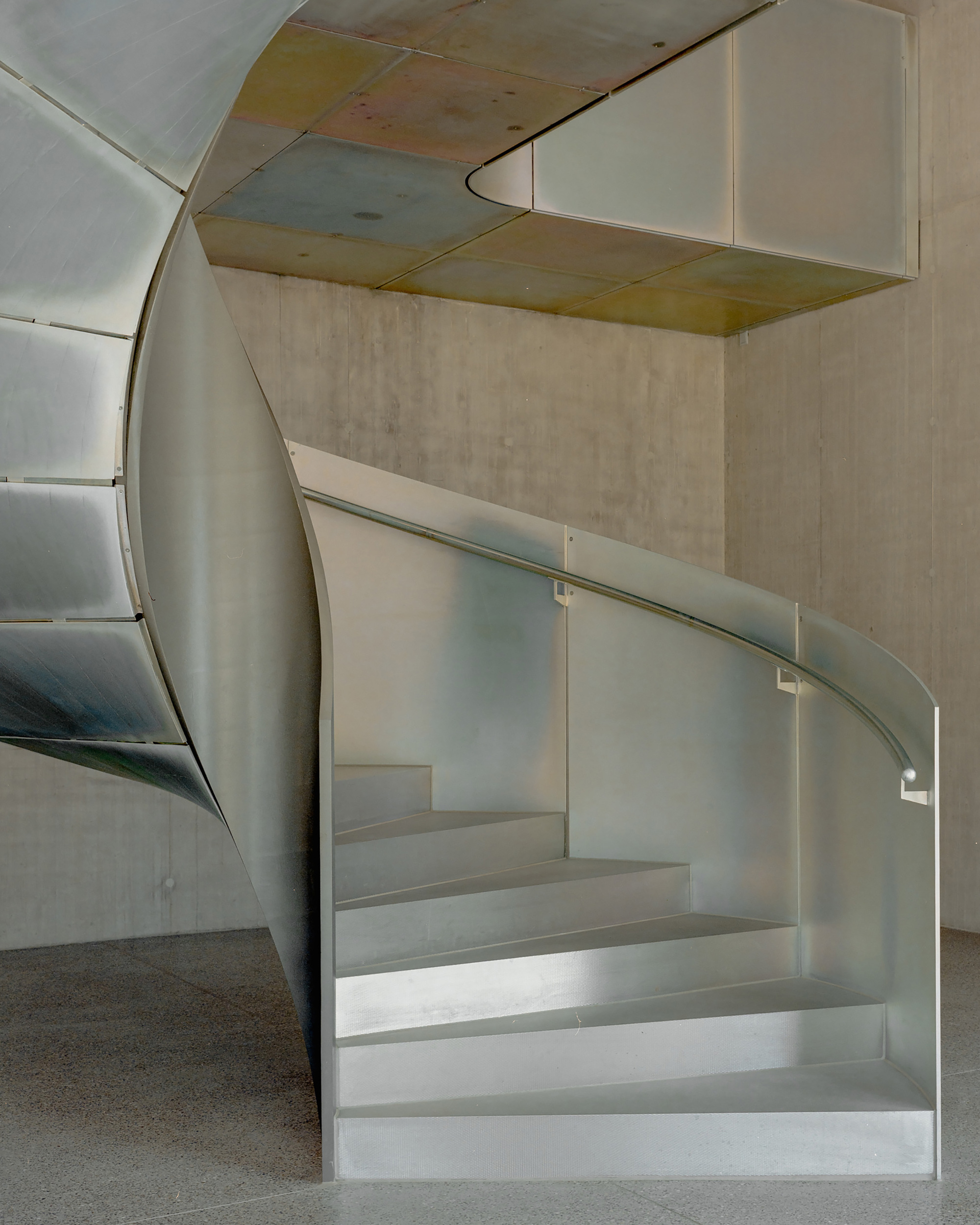 Spiral stair clad in sheet metal beneath a ceiling of oxidised metal panels, set against raw concrete walls