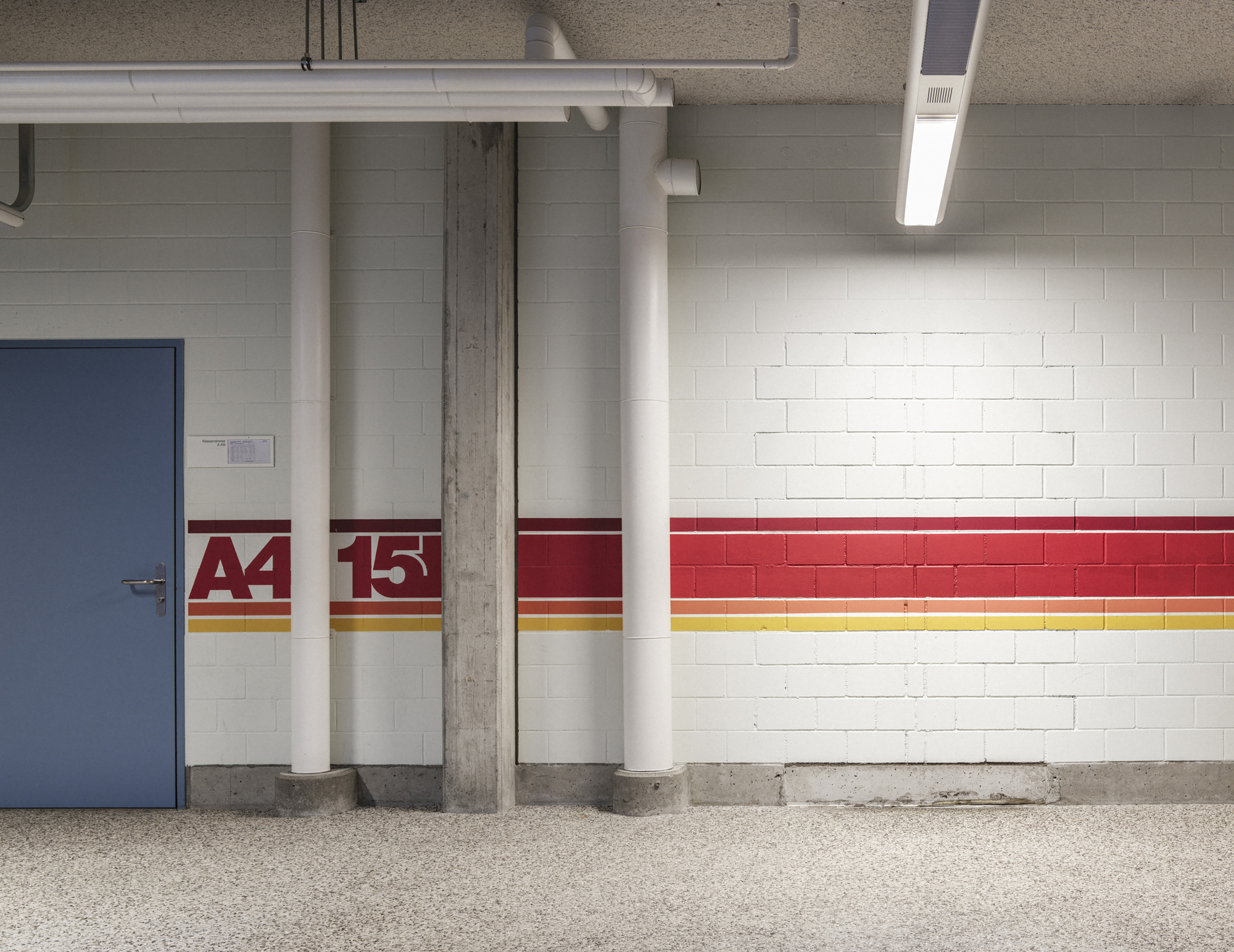 Whitewashed block wall with red and yellow stripes, a painted A4 15 marker, a blue steel door and white pipes under fluorescent light
