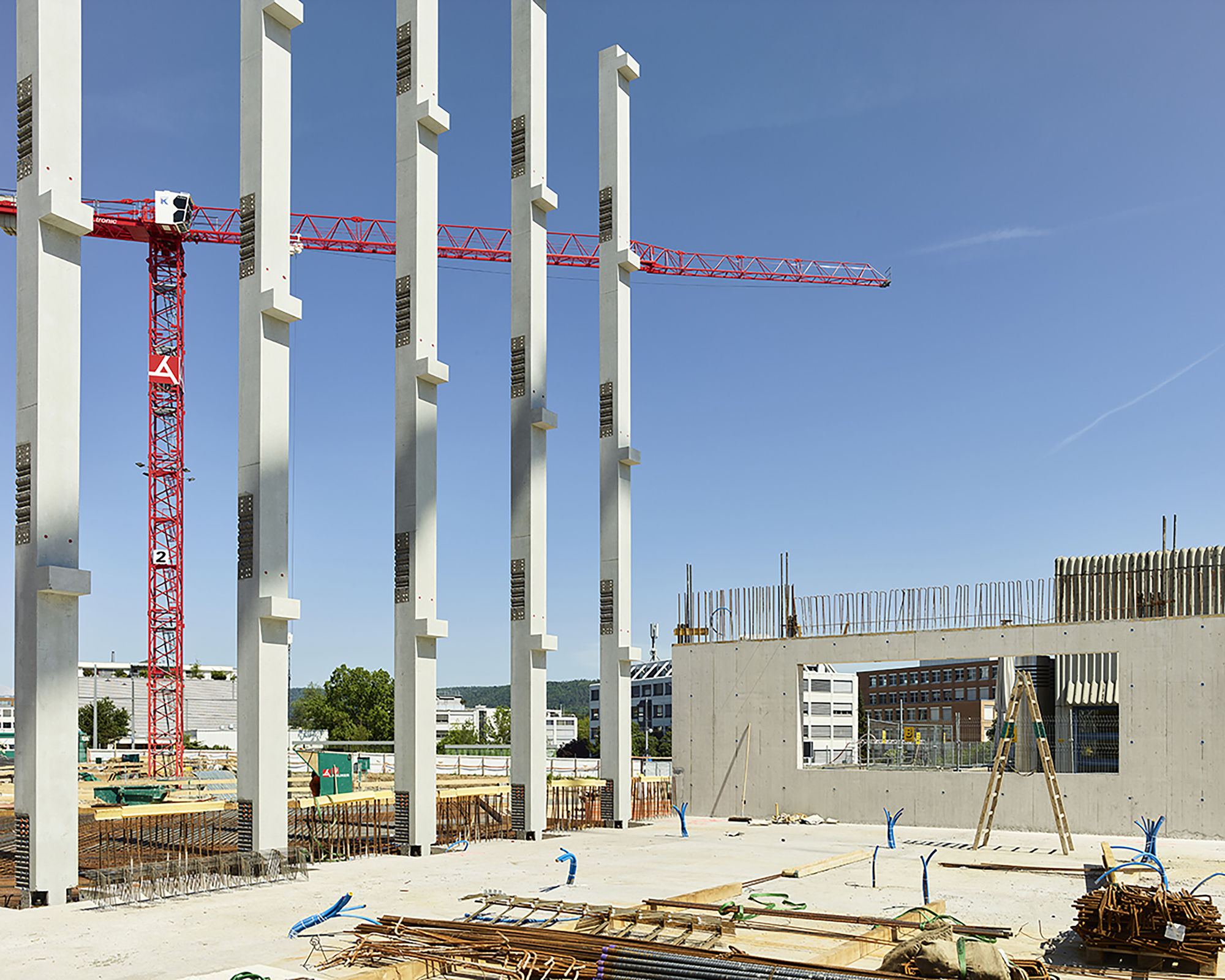 Row of precast concrete columns beside a red tower crane and a concrete wall with openings on a sunny construction site