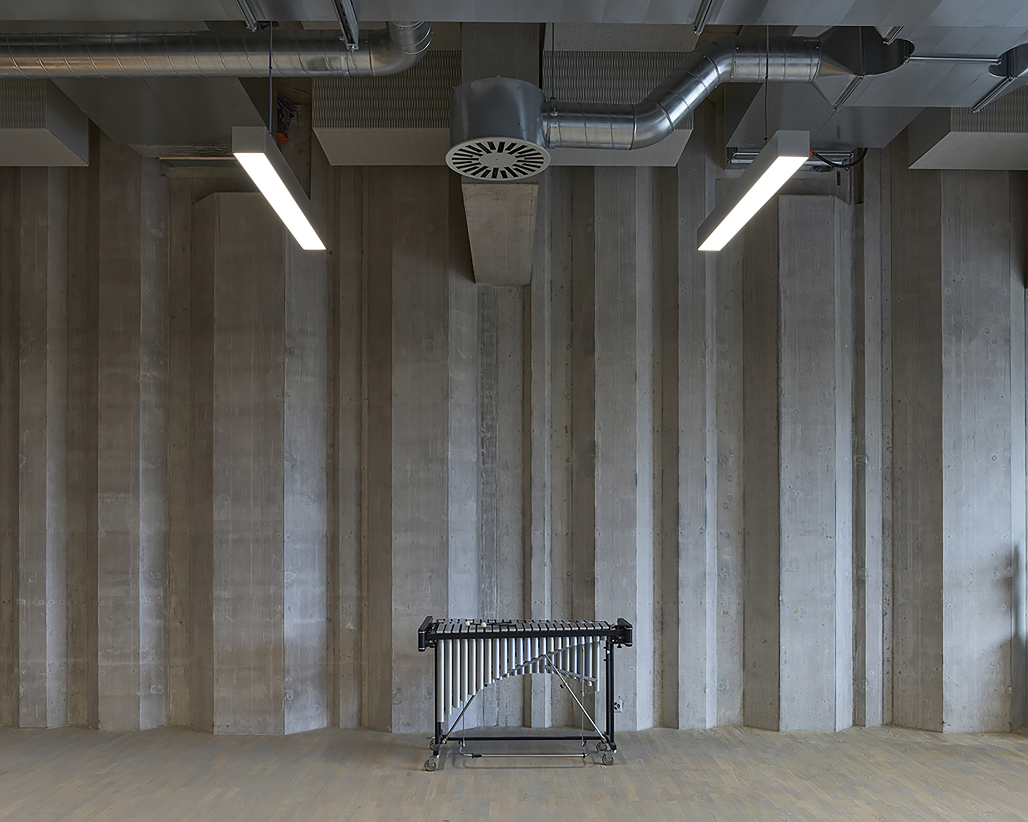 Board-formed concrete wall with exposed ducts and linear pendant lights above a vibraphone standing alone on a parquet floor