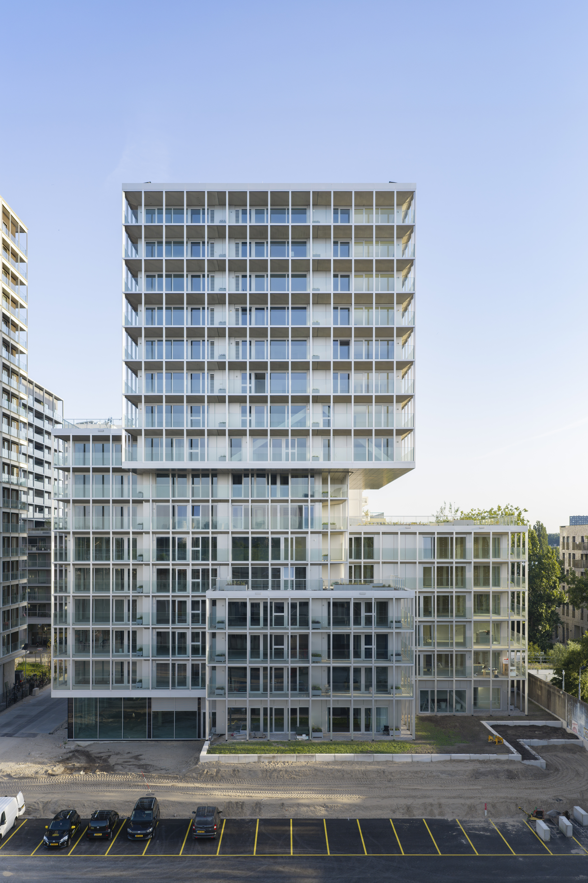 Stepped residential tower with glazed balcony grid rising above a gravel lot and marked parking bays at dusk
