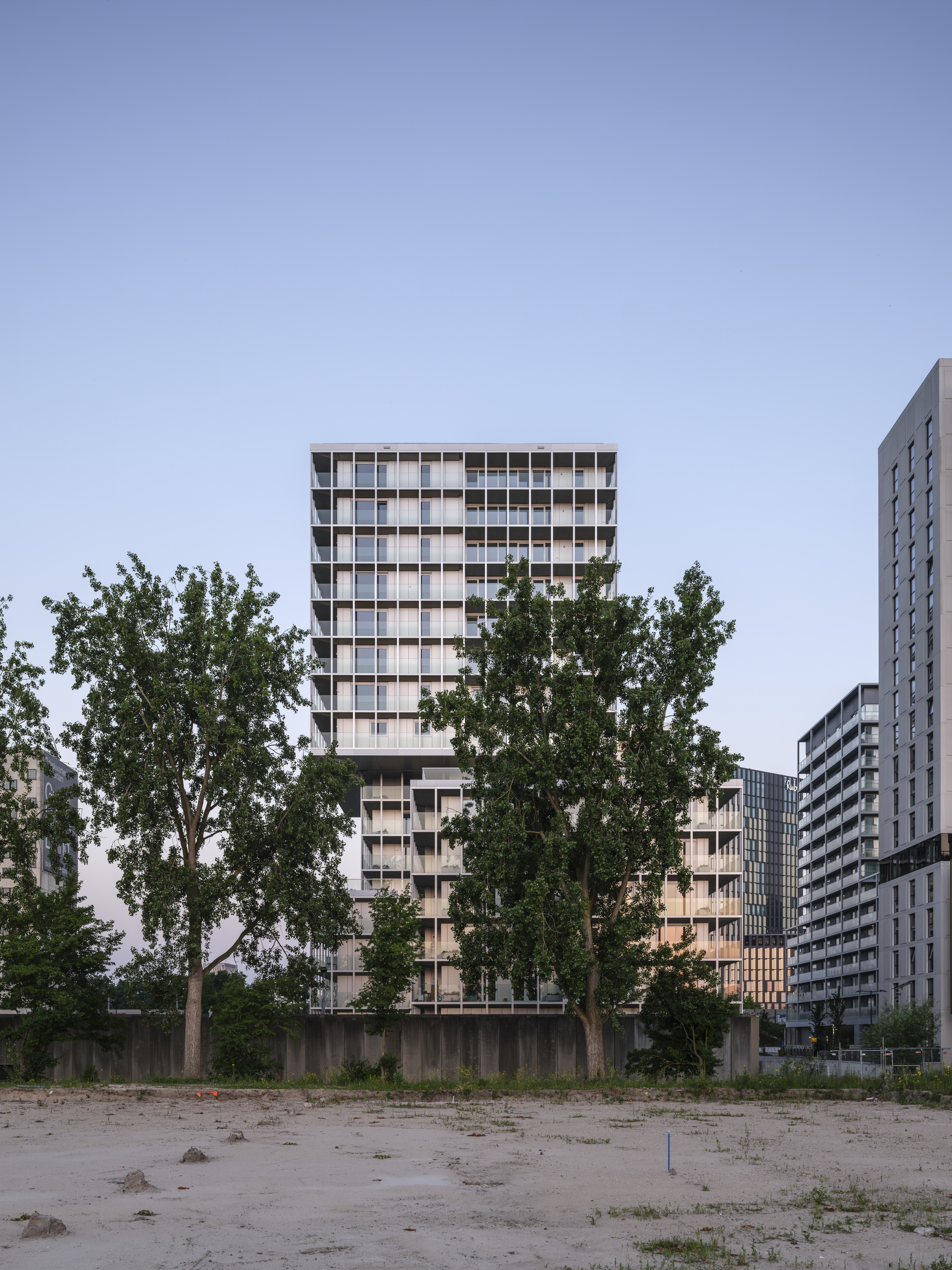 Tall gridded residential tower rising behind a concrete wall and two poplar trees across a bare sandy lot at dusk