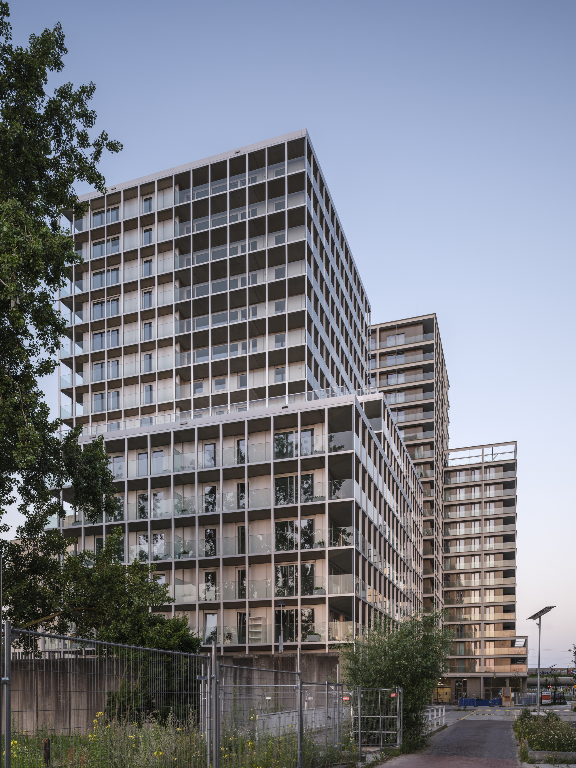 Stepped tower volumes with deep balcony grid seen from a brick-paved lane past a construction fence in evening light