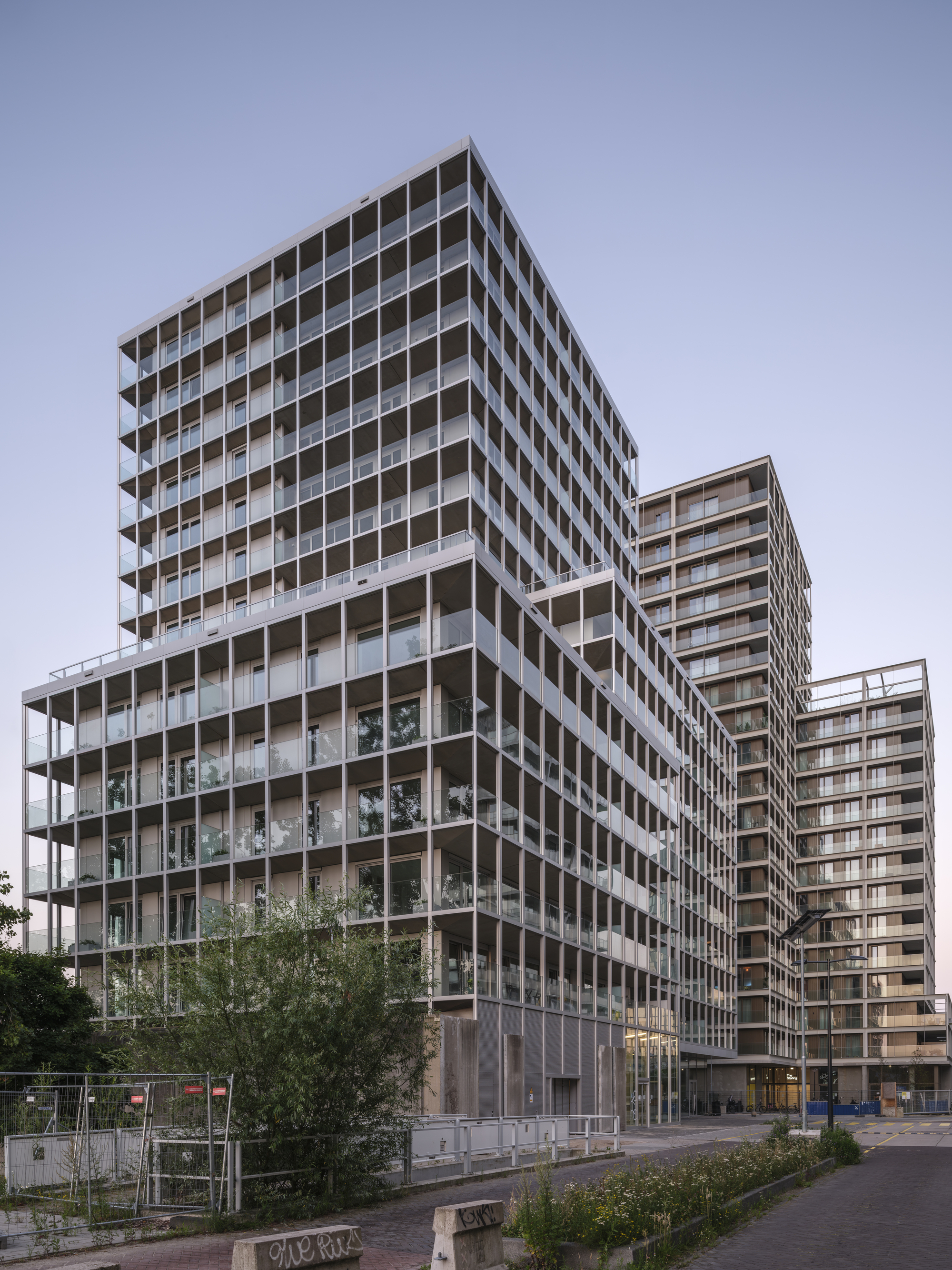 Corner view of stacked towers with open balcony frames, a young tree and graffitied bollards in the foreground at dusk