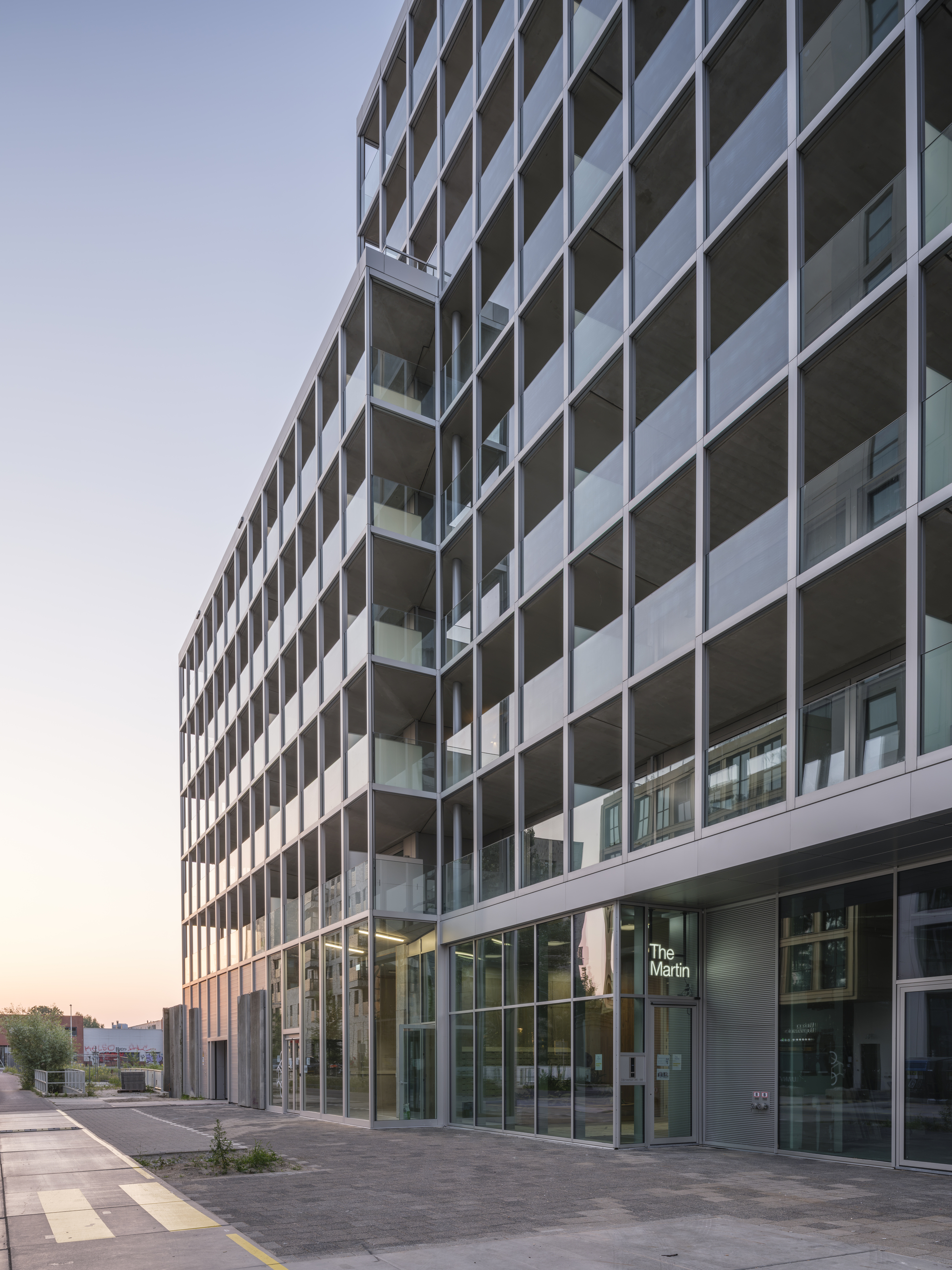 Ground-floor entrance with glass lobby beneath rows of recessed balconies, with a paved sidewalk and pink dusk sky at left