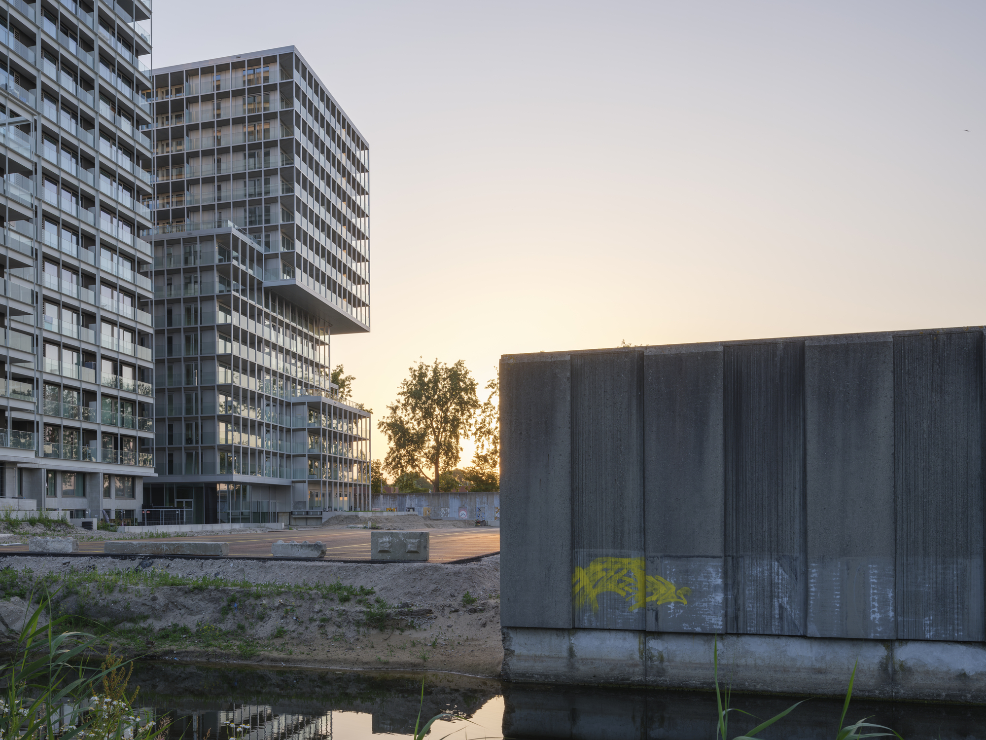 Gridded tower blocks beyond a canal edge and a graffiti-marked concrete wall, lit by a low orange sunset