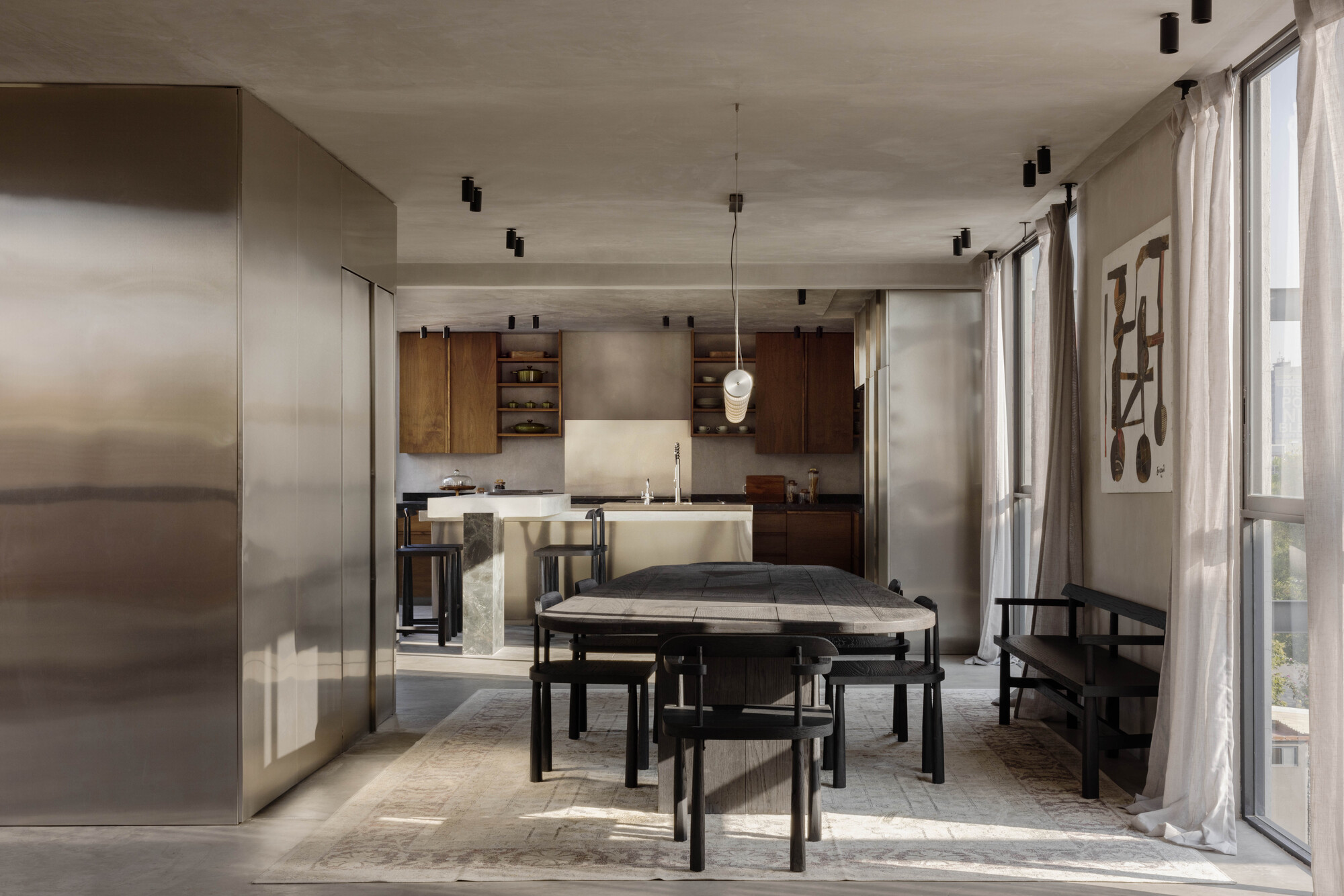 Dining table and black chairs before an open kitchen with walnut cabinets, framed by a steel-clad volume and sheer curtains in low sun
