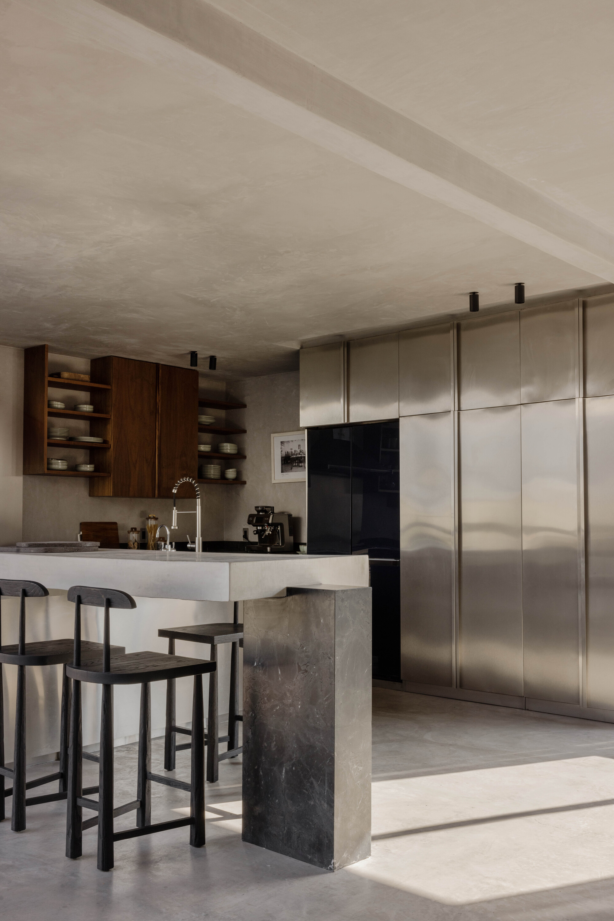 Kitchen island with a dark stone end panel and black stools beside a wall of brushed steel cabinets, with a patch of sunlight on the floor