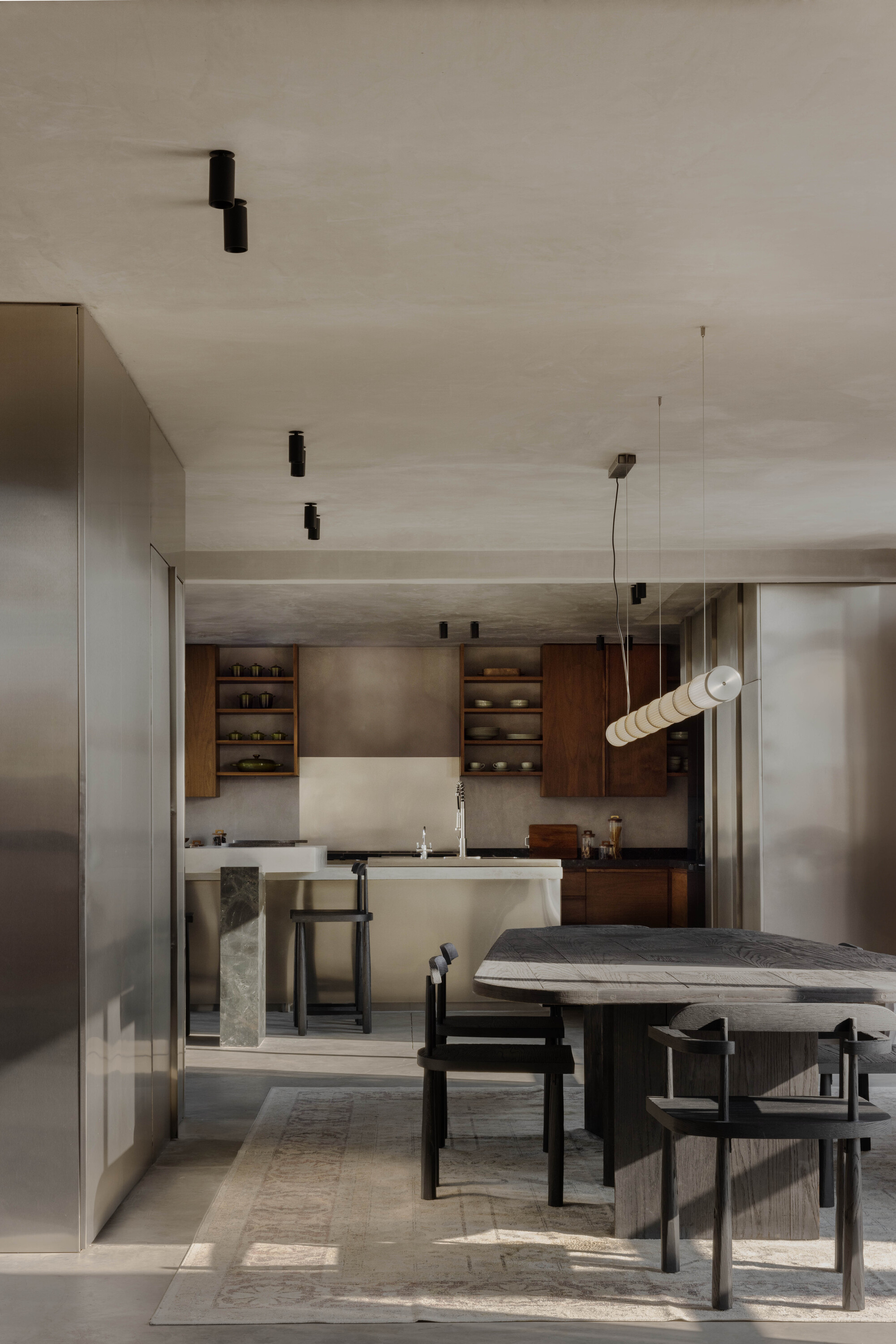 View past a steel-clad wall to the kitchen island, walnut shelves and a dark dining table, with a linear pendant lamp and sunlit rug